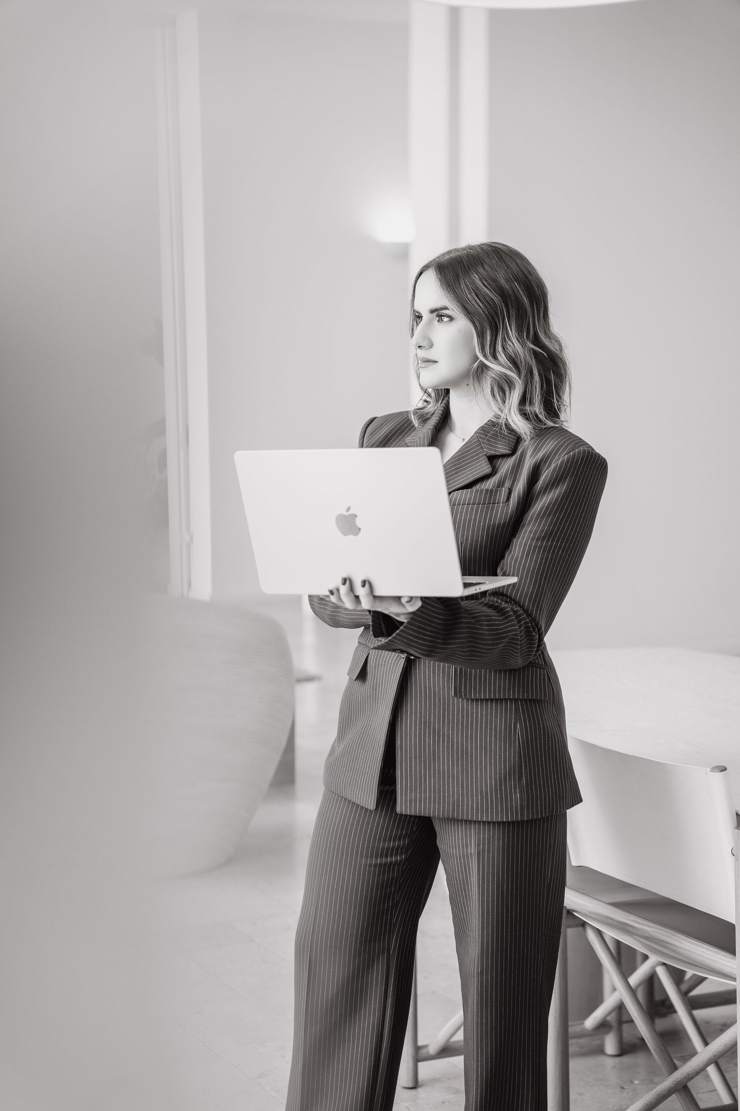 Black and white photo of a professional woman in a pinstripe suit holding an Apple MacBook laptop in a modern office.