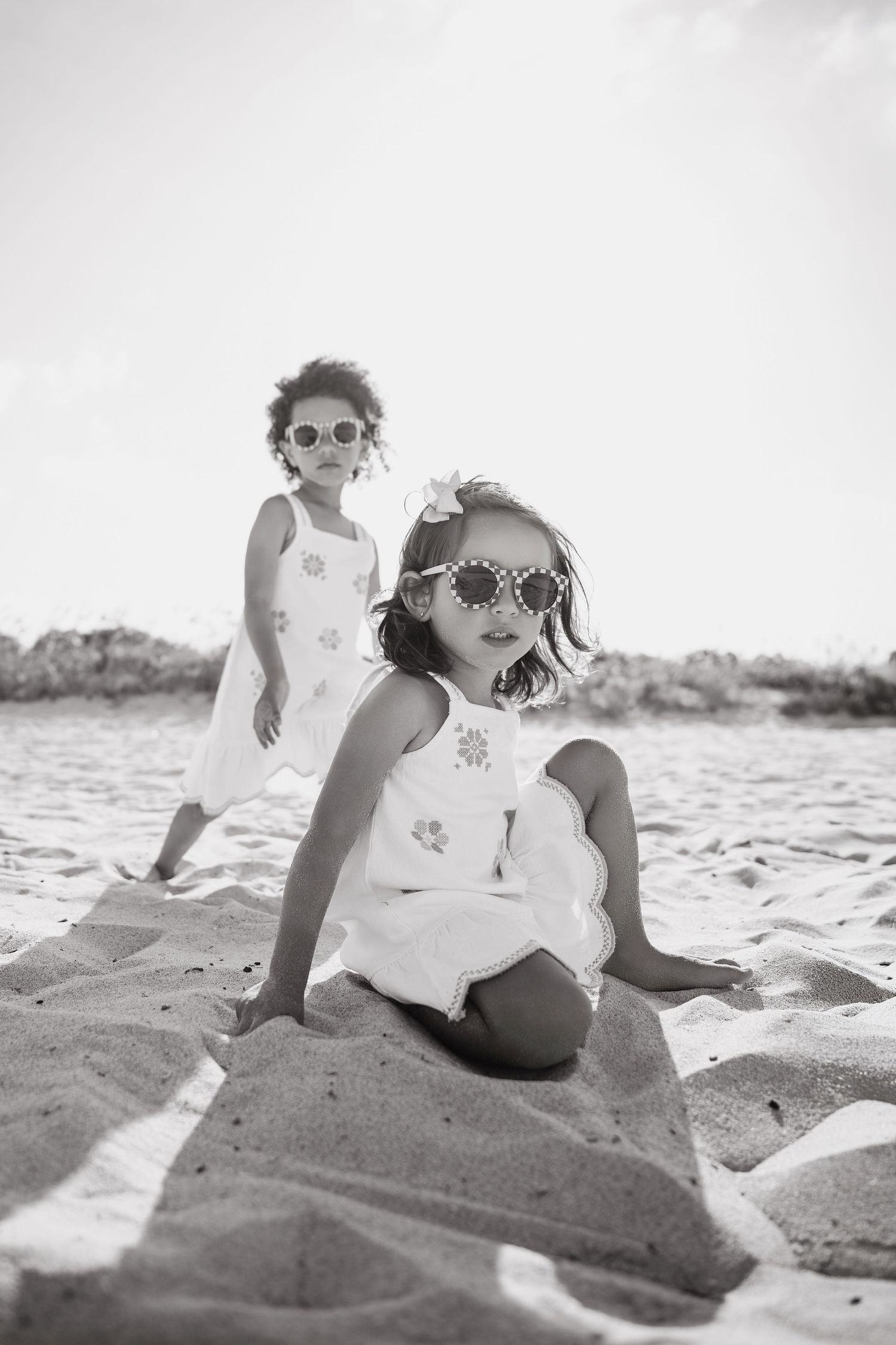 Two young girls in white dresses and flower sunglasses play on a sandy beach in a black and white photo.