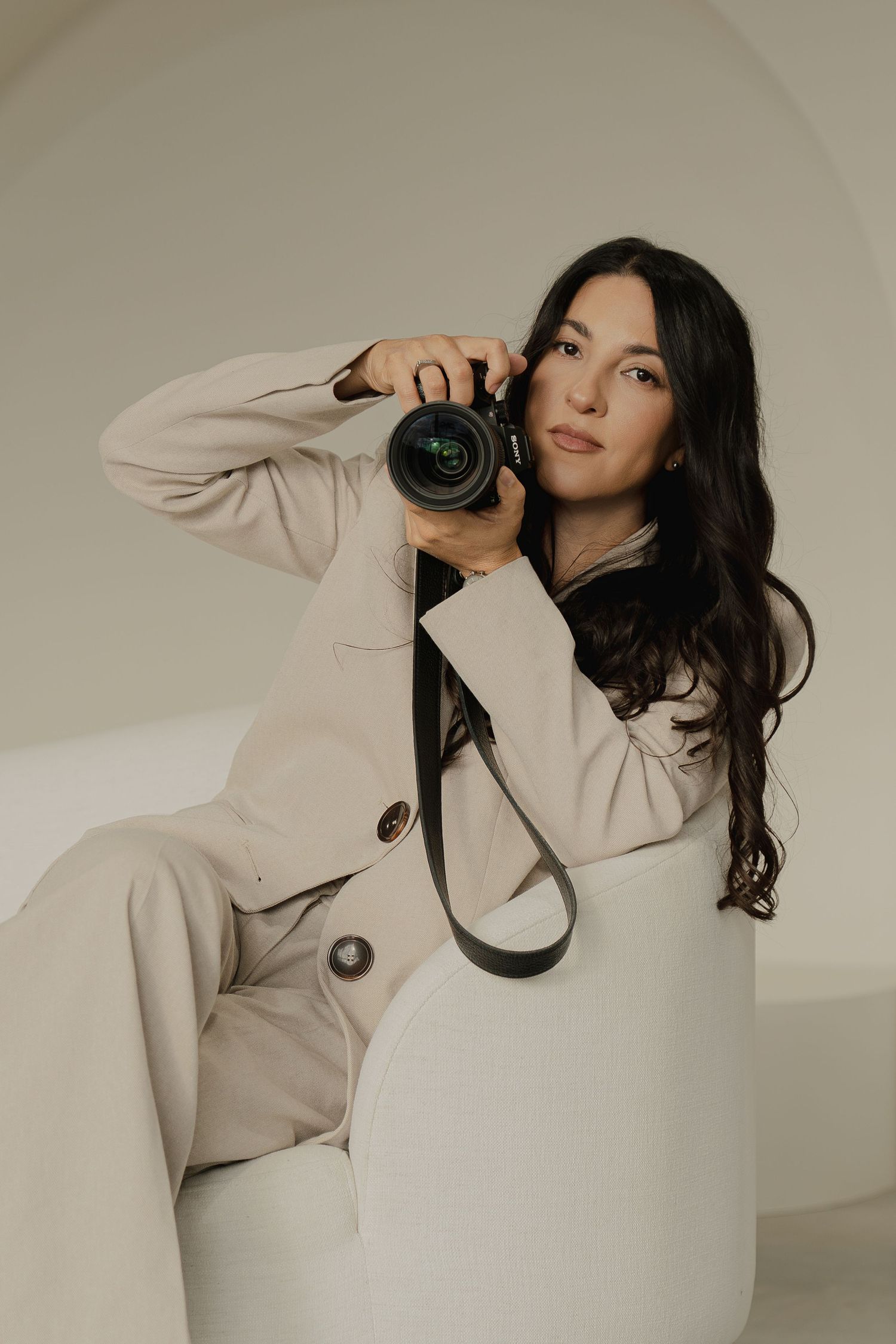 Woman in beige suit holding a DSLR camera while seated in a modern white chair against a neutral background.