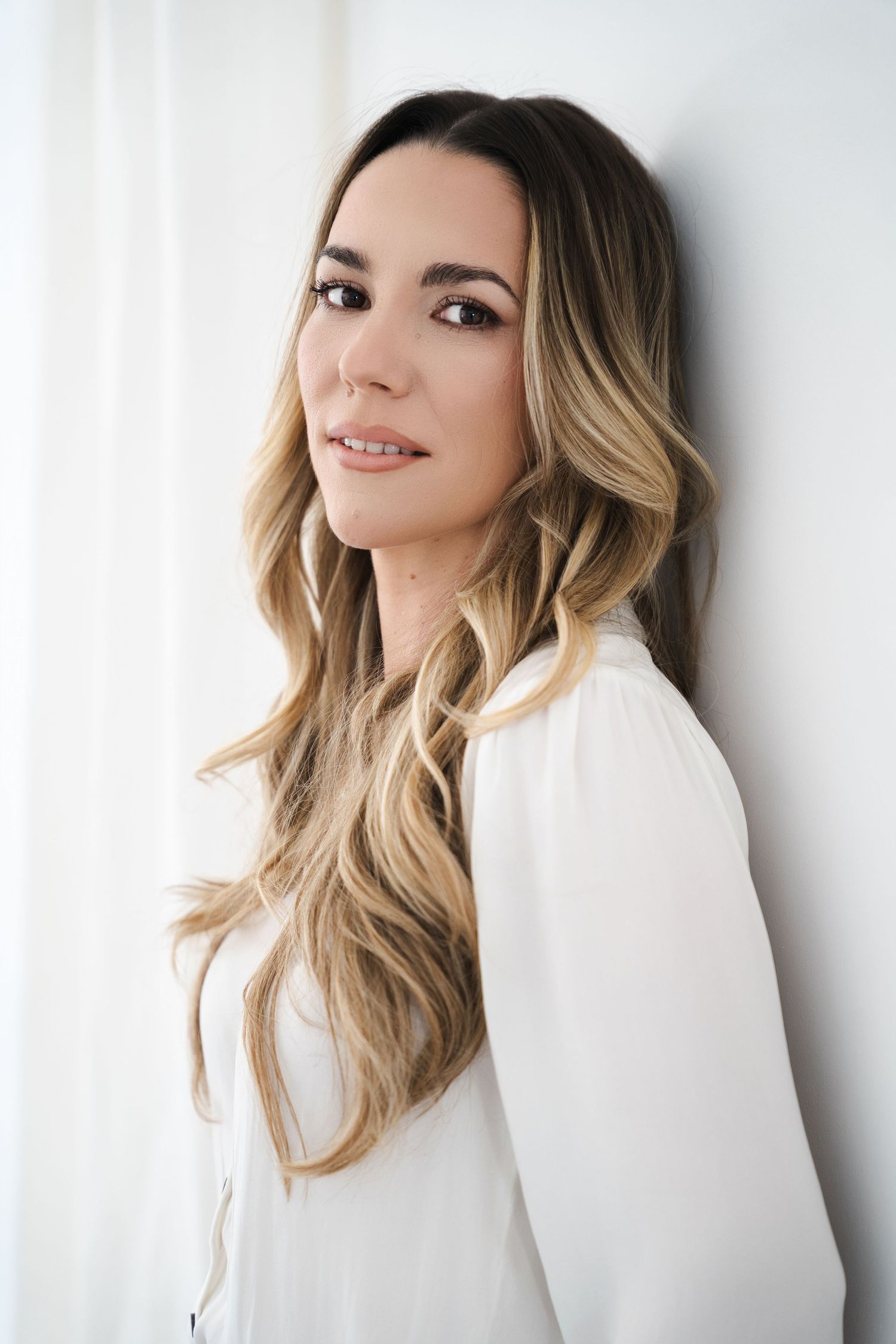 Young woman with long blonde ombre hair wearing a white blouse, posing against a white wall.