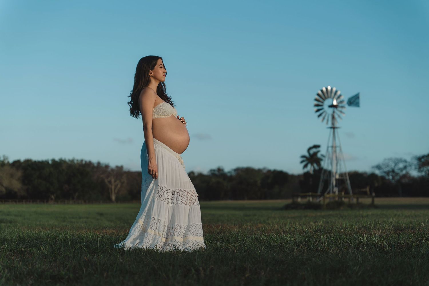 A pregnant woman in a white lace dress stands in a rural field with a windmill silhouetted against the blue sky.
