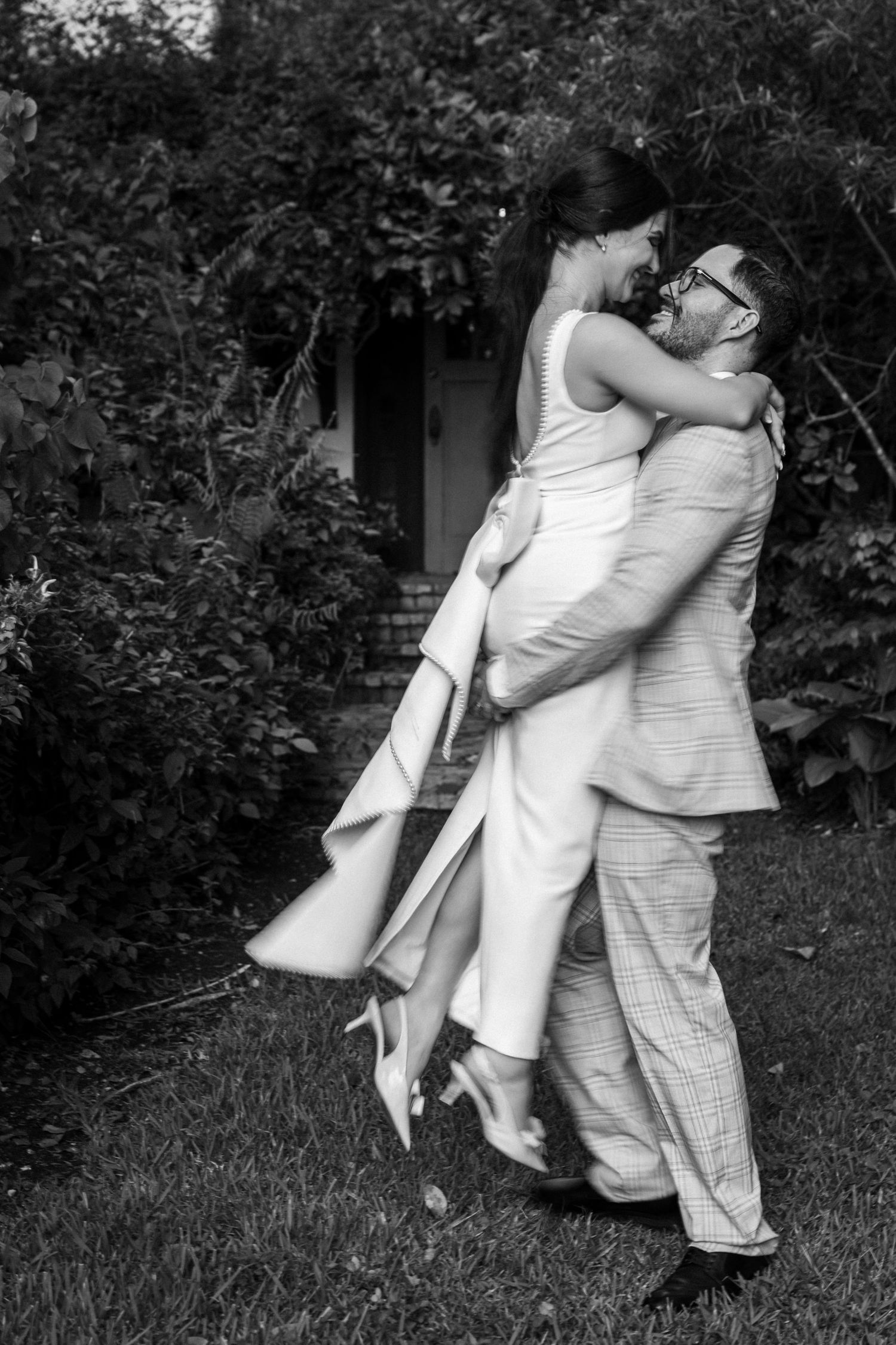 Black and white photo of a groom lifting his bride in a garden, sharing a romantic moment.