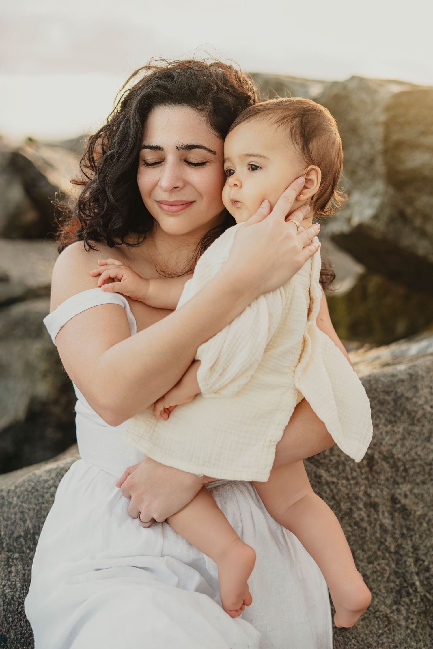 Sweet maternal moments captured on rocky coastal terrain in white outfits.