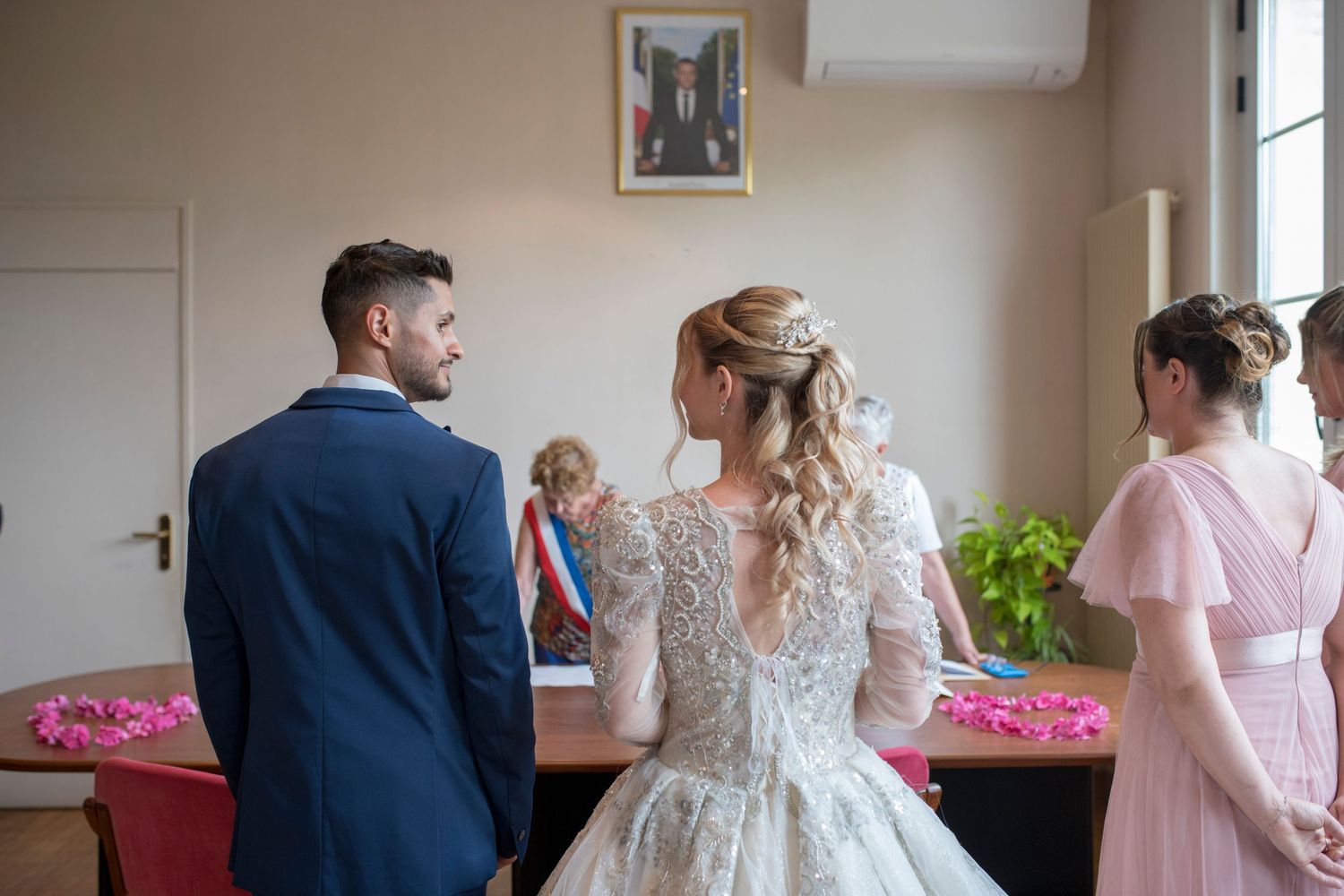 Une mariée en robe de mariée en dentelle se tient à une table de réception avec des invités en tenue de soirée près d'un mur avec une photo