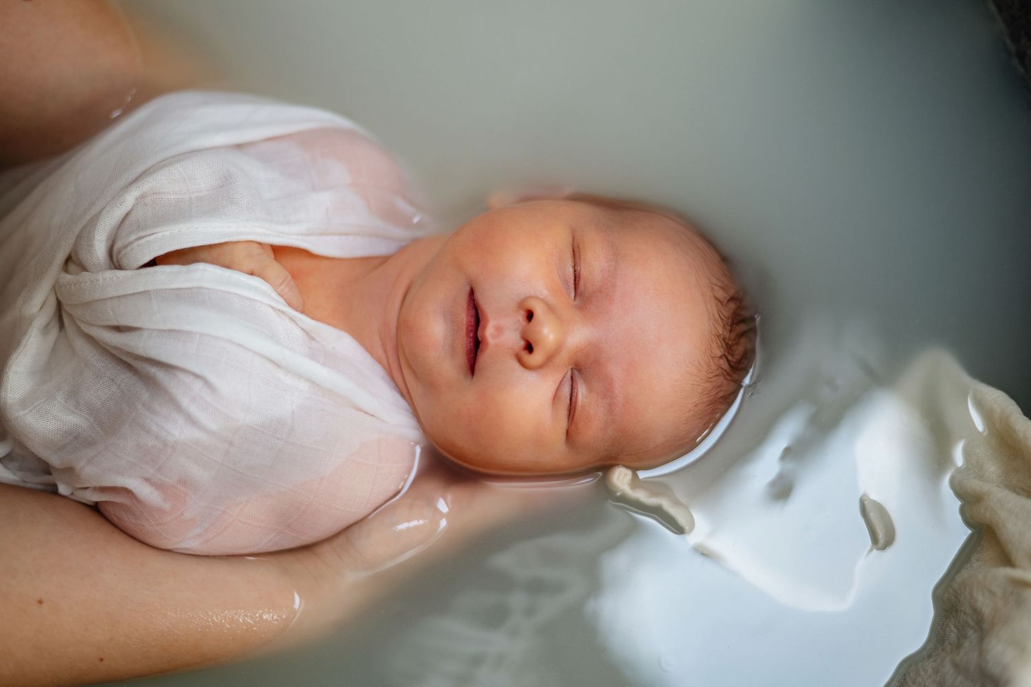 Séance photo bain enveloppé, bébé endormi paisiblement, vêtu de vêtements blancs, repose sur une literie douce à la lumière naturelle.