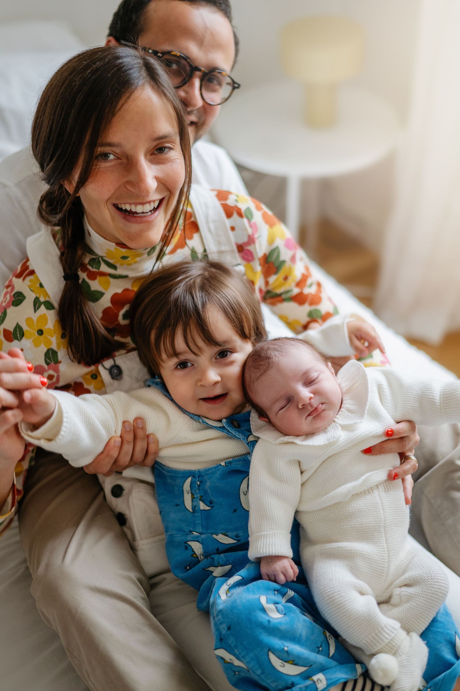 Famille heureuse assise ensemble sur le canapé, un tout-petit portant une salopette bleue et un nouveau-né en tenue blanche.