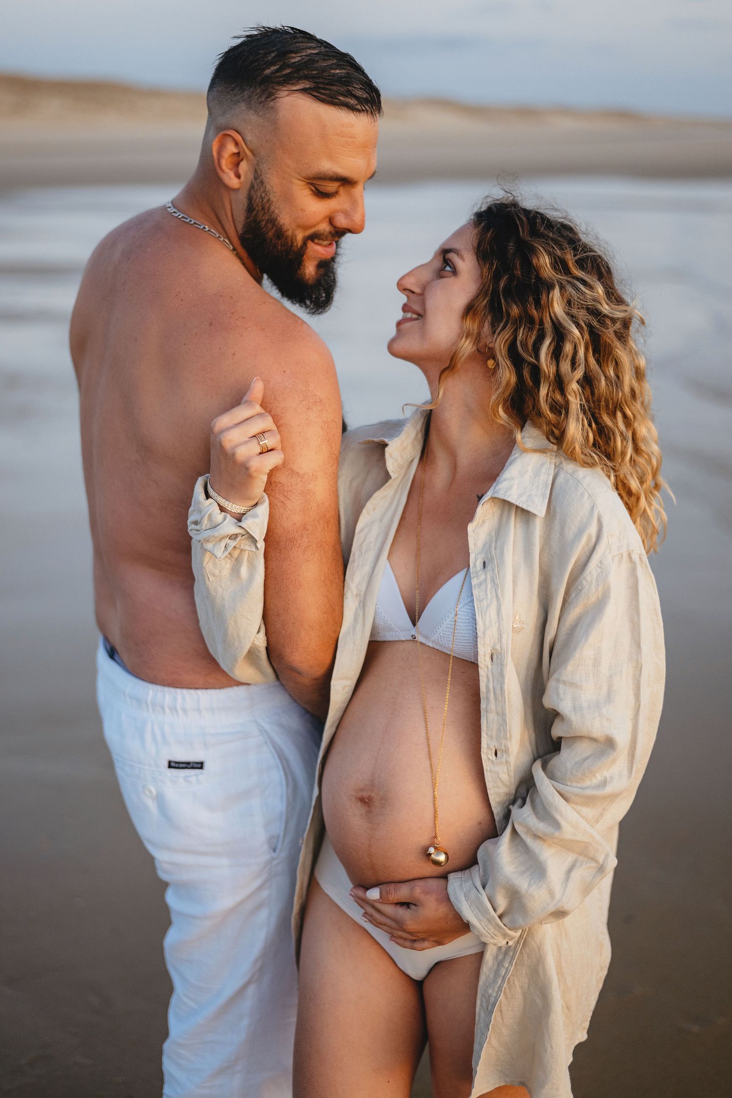 Photo de maternité romantique d'un couple s'embrassant sur la plage au coucher du soleil en tenue de plage décontractée.
