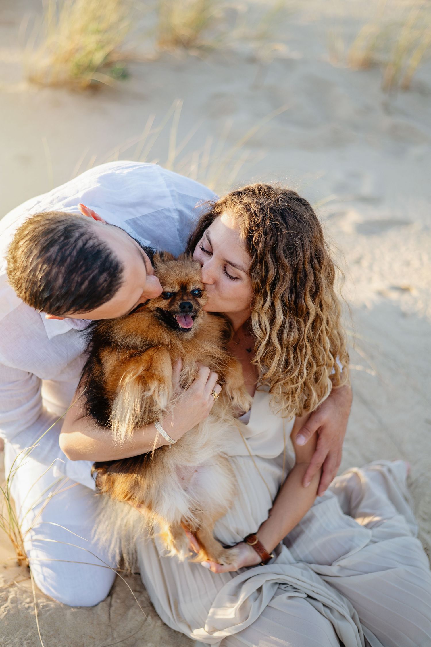 Un couple en tenue blanche partage un moment de tendresse avec leur petit chien tout doux à la plage pendant l'heure dorée.