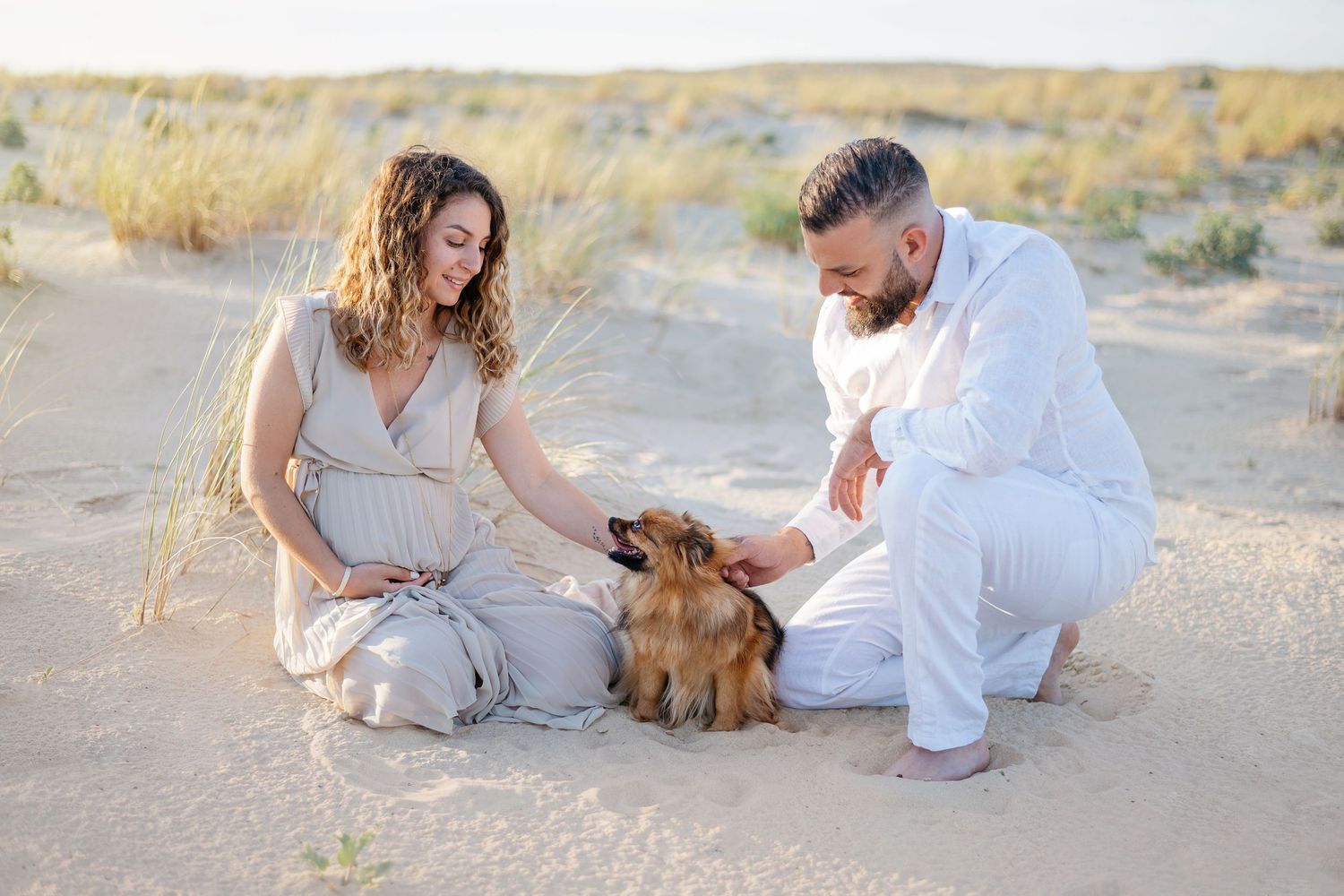 Couple en tenue blanche assis sur un sol sablonneux avec un petit chien marron dans un paysage désertique.