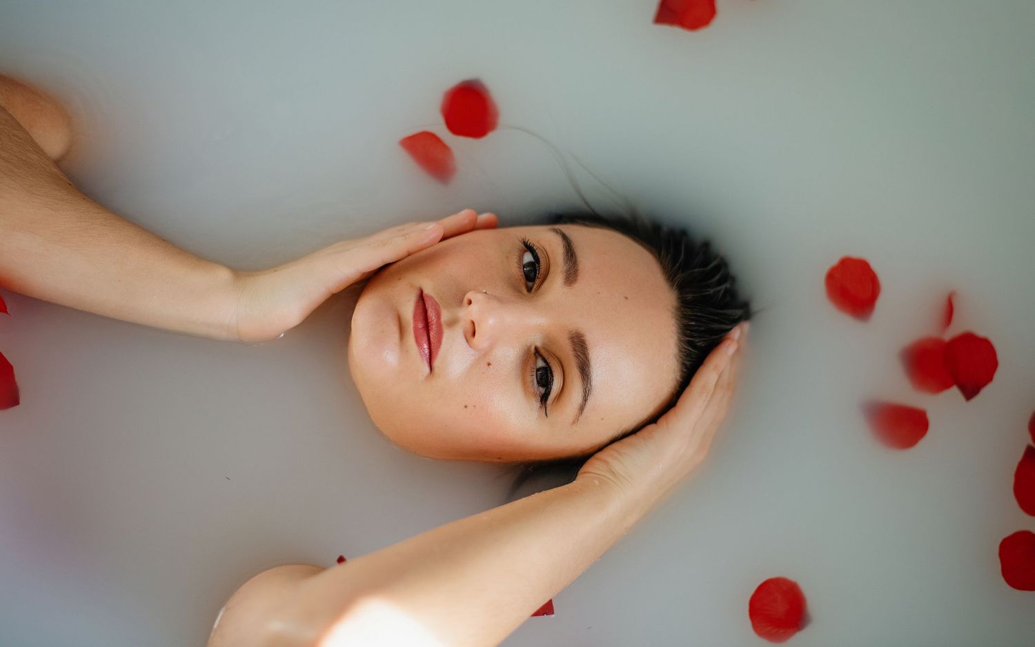 Portrait artistique de baignoire avec des pétales de roses rouges dispersés sur une surface d'eau blanche laiteuse.