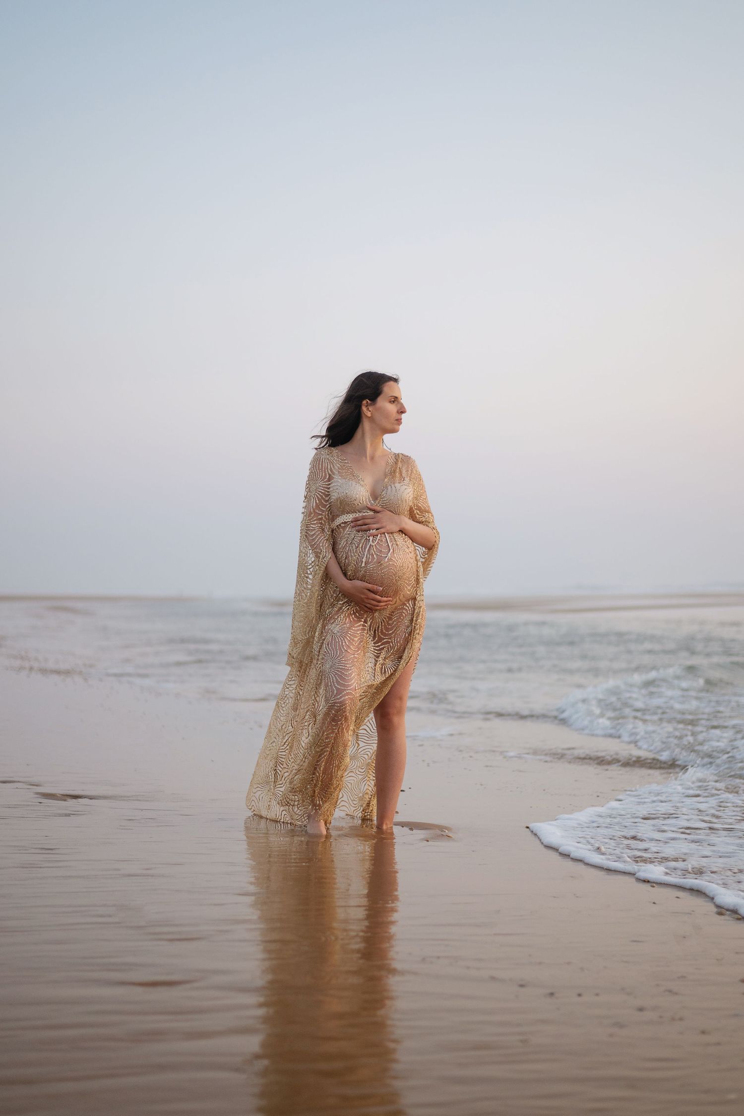 Une femme enceinte vêtue d'une robe fluide se tient au bord de l'océan au coucher du soleil, les vagues clapotant doucement à ses pieds.