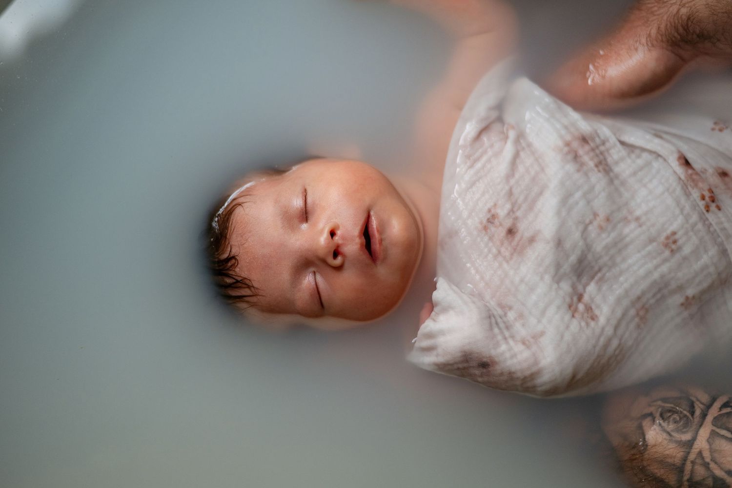 A newborn baby sleeps peacefully while swaddled in a white textured blanket.