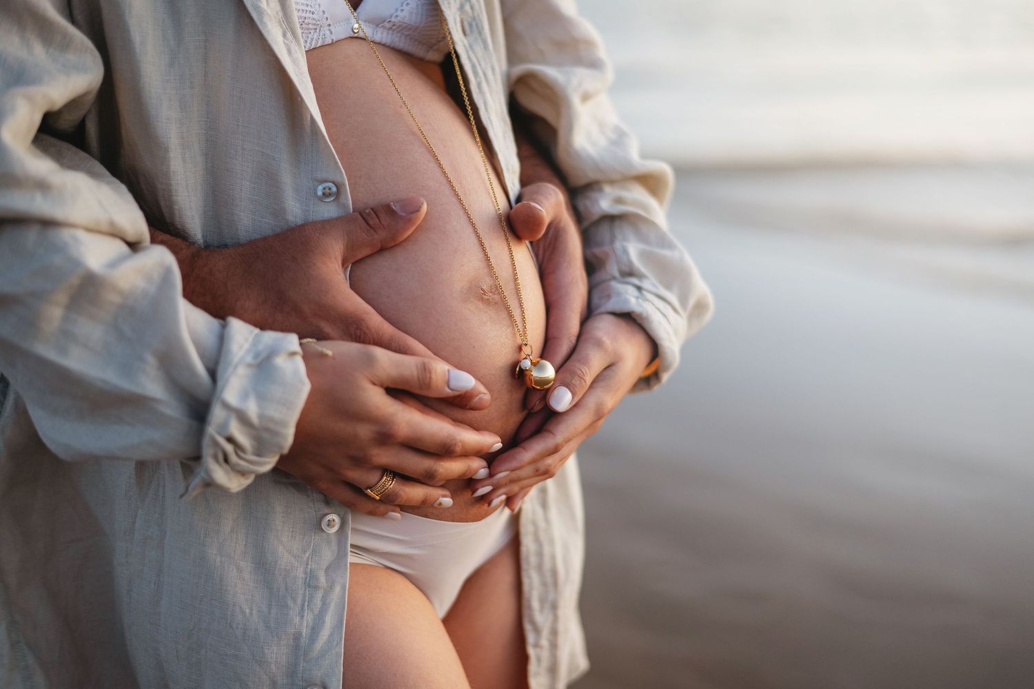 Vue rapprochée d'un ventre de femme enceinte tenu par des mains portant une chemise de couleur claire au bord d'un front de mer tranquille