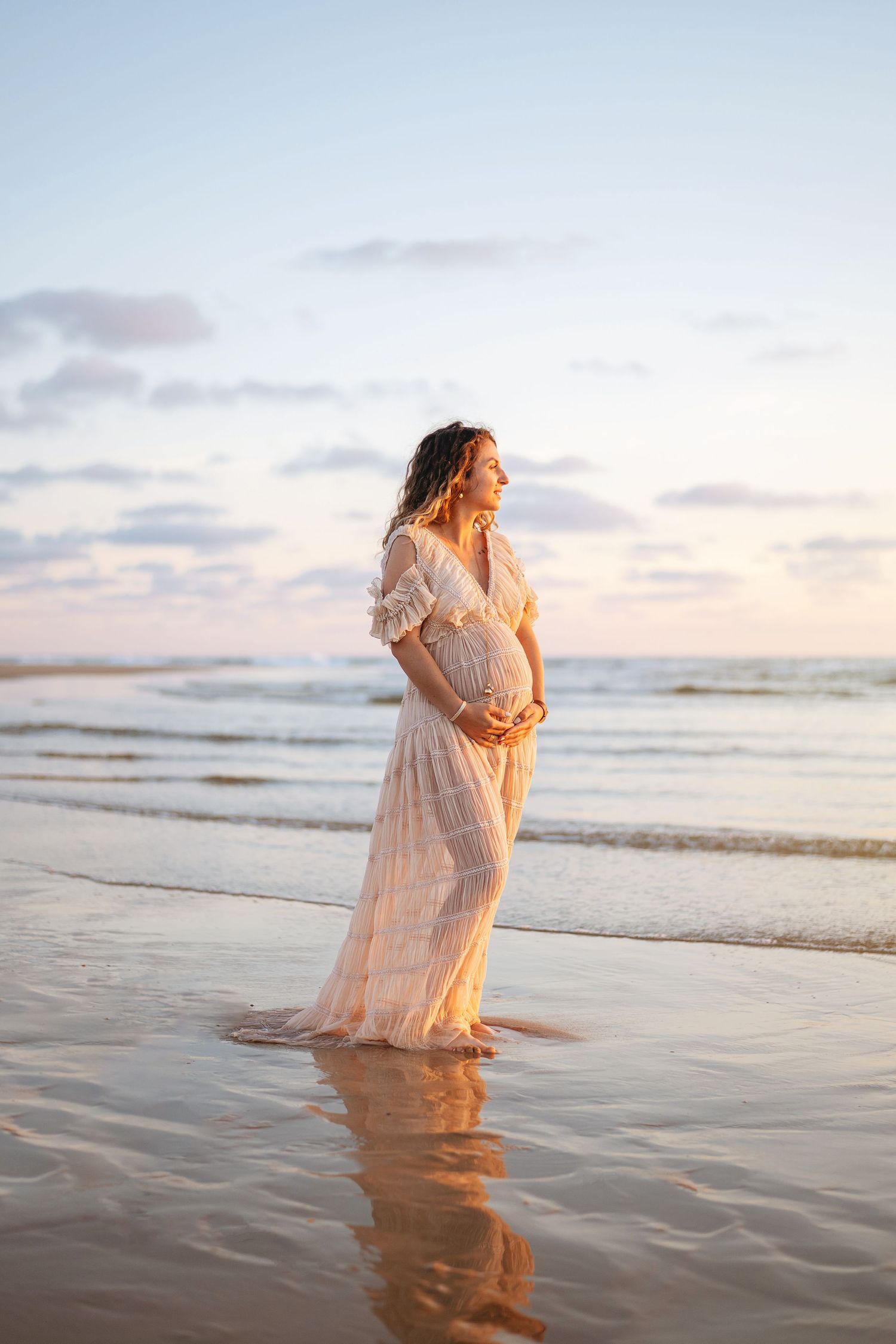 Une femme enceinte vêtue d'une robe blanche fluide se tient sur une plage au coucher du soleil, les vagues clapotant doucement à ses pieds.