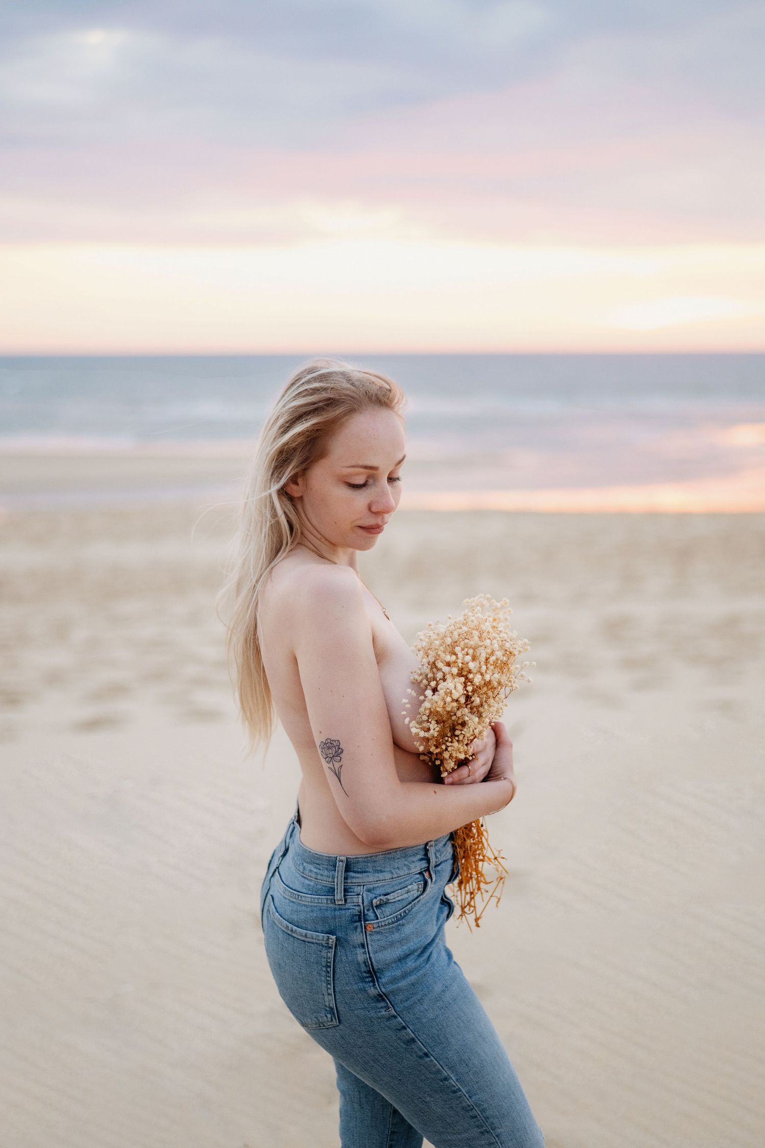 Une personne en jean tenant des fleurs séchées se tient sur la plage au coucher du soleil avec un ciel pastel doux en arrière-plan.
