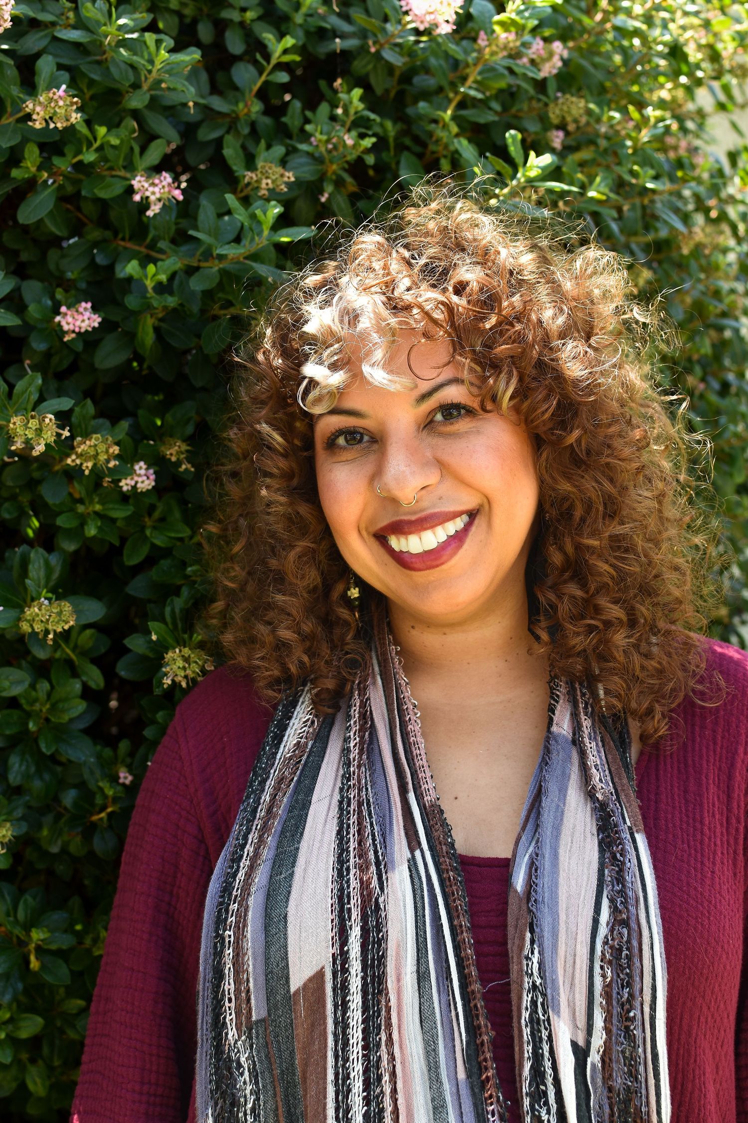 A smiling person with curly hair wearing a burgundy sweater and striped scarf stands in front of flowering shrubs.