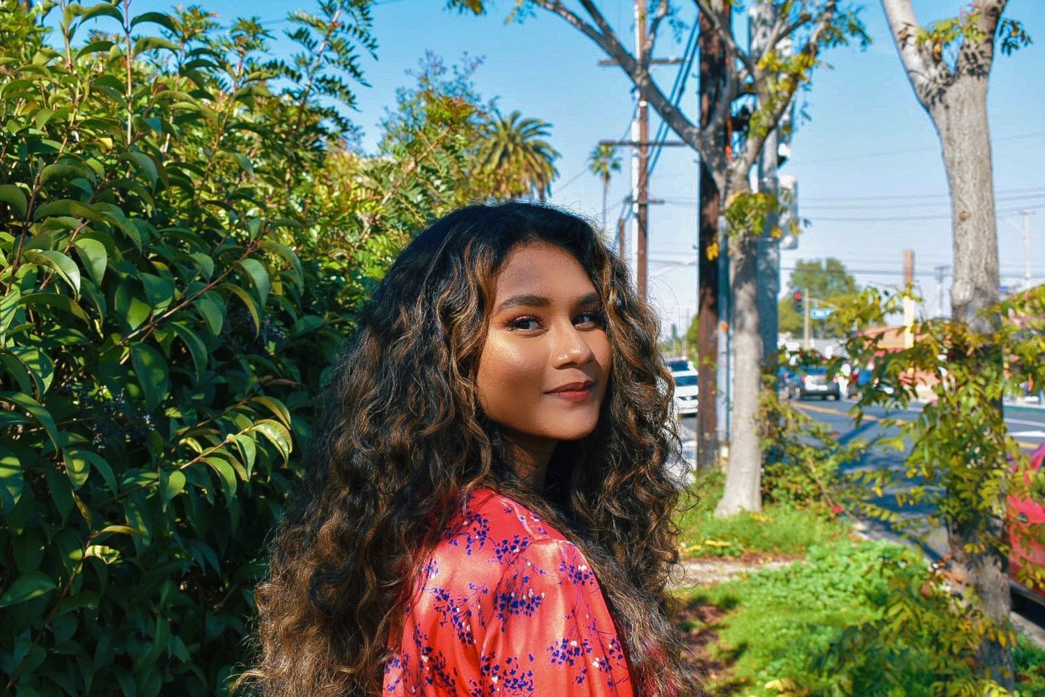 Person with long wavy hair poses outdoors in a pink floral dress against lush greenery.