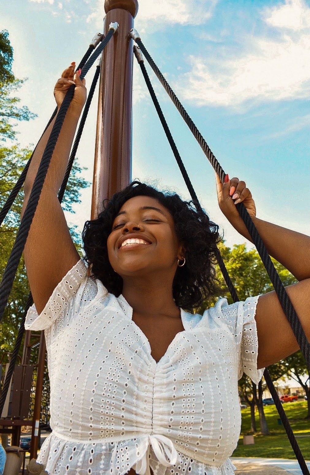 Joyful moment captured on playground swing wearing white eyelet top against blue sky.