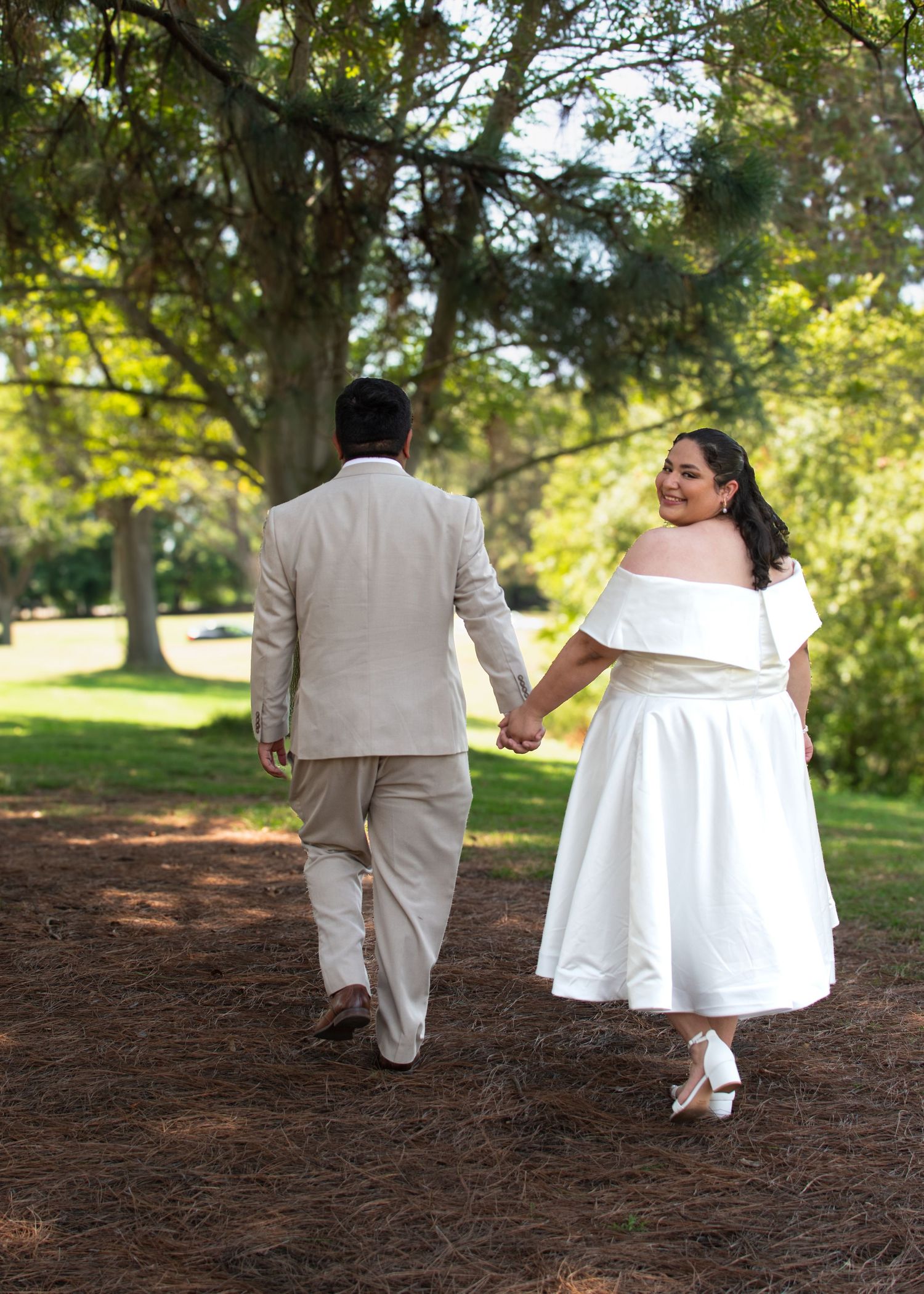 A newlywed couple walks hand in hand down a wooded path, with the bride in white and groom in beige.