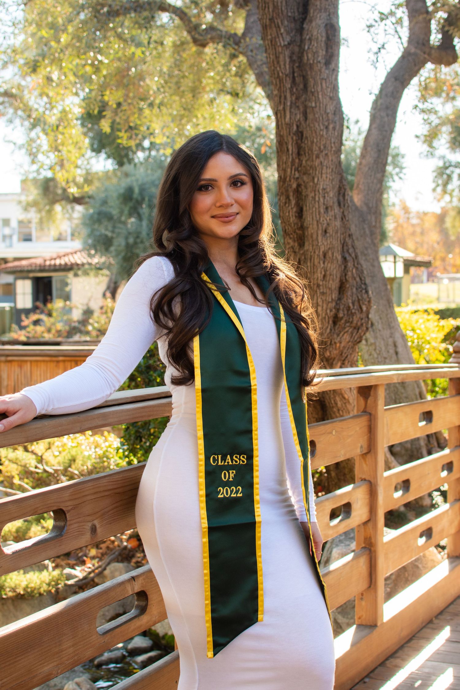 Graduate in form-fitting white dress and dark green graduation sash poses on wooden deck with trees in background.