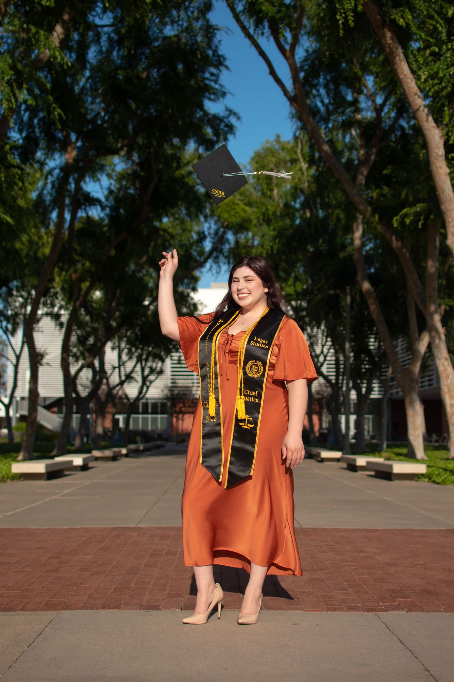 Graduate in orange cap and gown celebrates on campus pathway surrounded by trees.