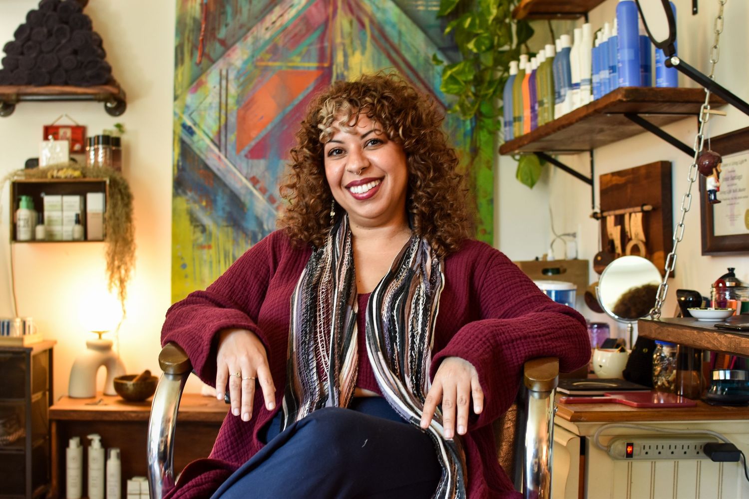 Someone with curly hair sits comfortably in a chair at a cozy, eclectic home space filled with decor and furnishings.