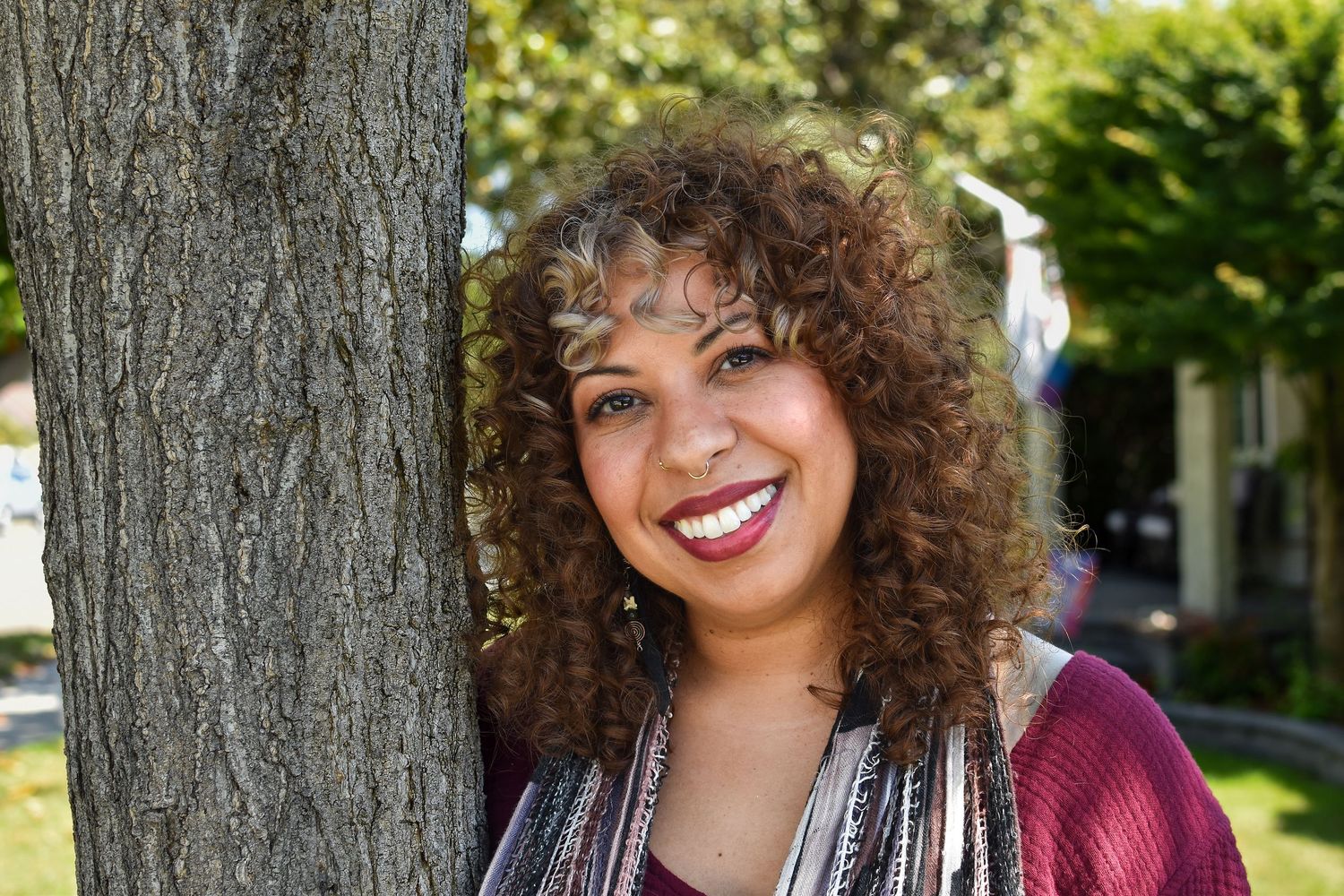 A person with curly hair and a bright smile leans against a tree trunk on a sunny day outdoors.