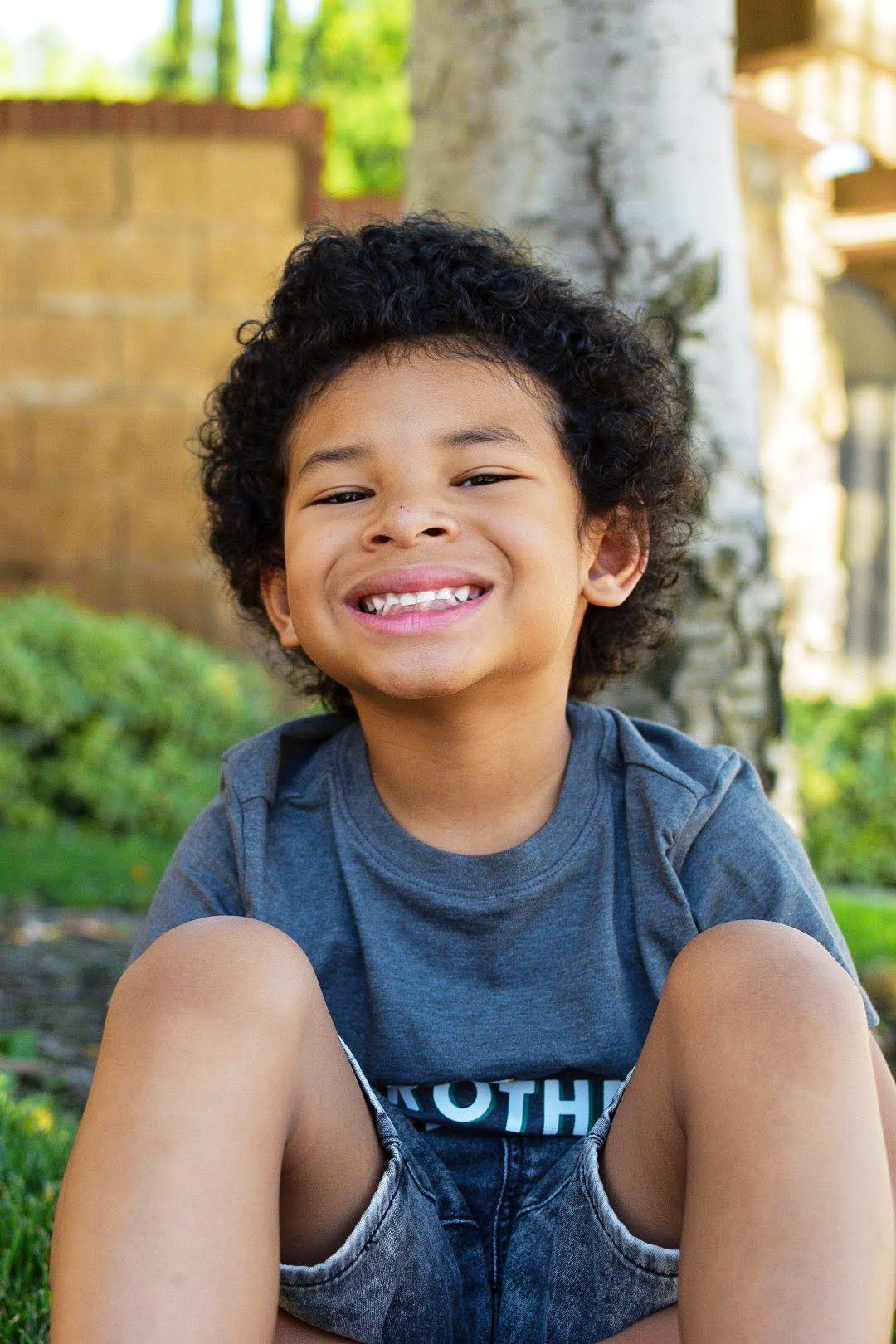 Child in casual attire sits outdoors while smiling brightly near a tree.