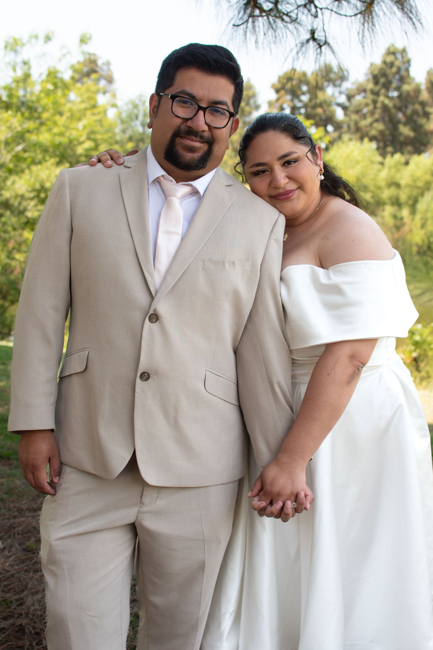 A newlywed couple in white and beige attire pose outdoors holding hands in a natural setting on their wedding day.