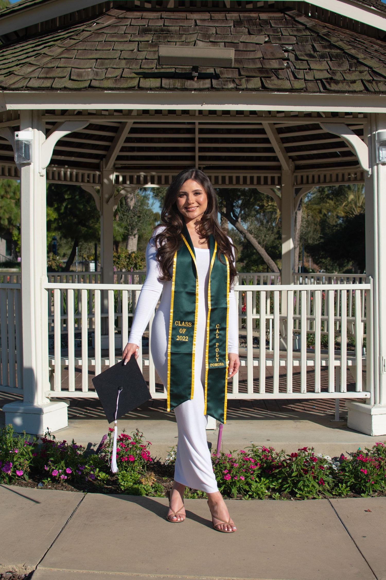 Graduate in white dress and green graduation stole poses playfully in outdoor gazebo setting.