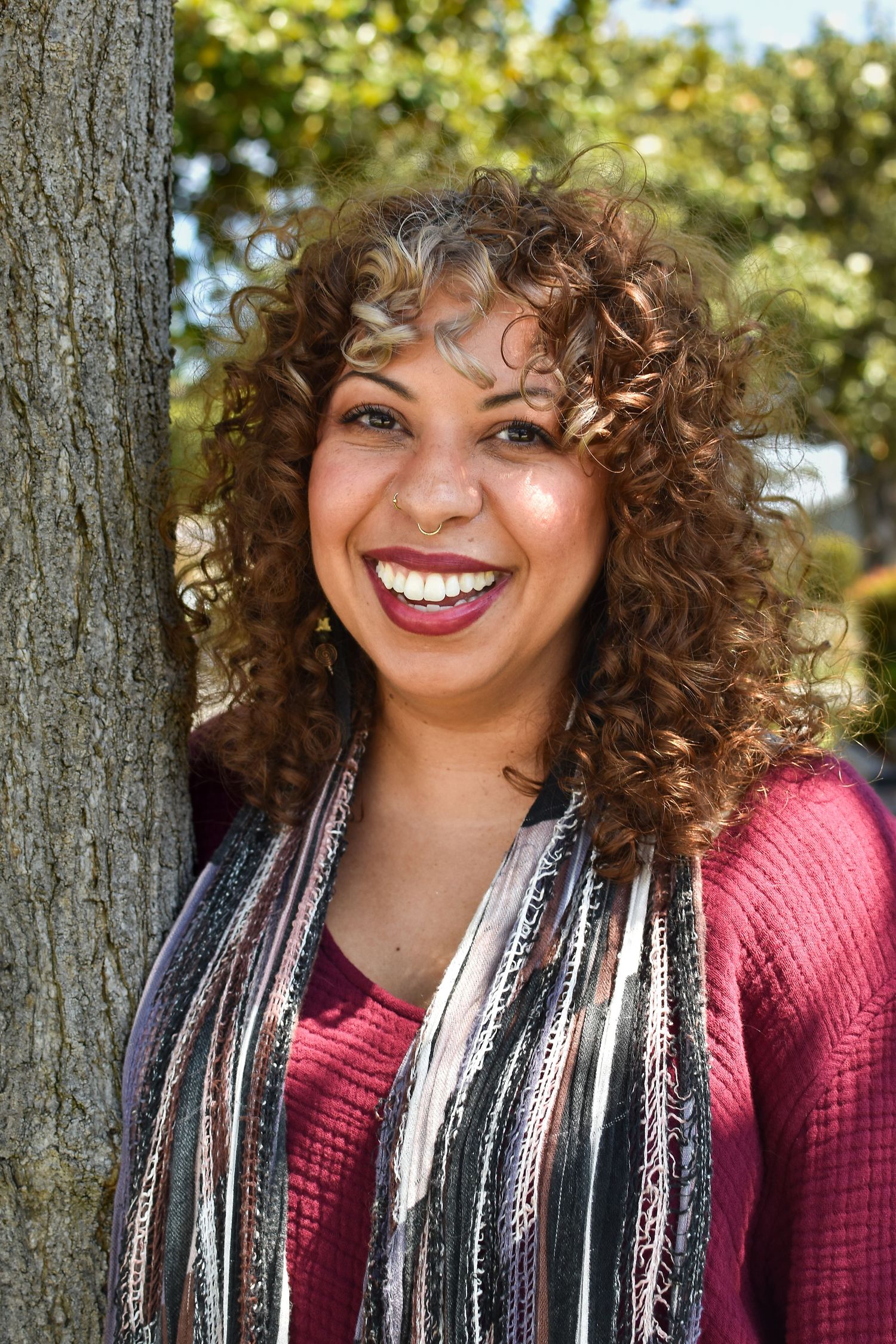 Portrait of a person in burgundy top and striped scarf against green foliage with a joyful expression.