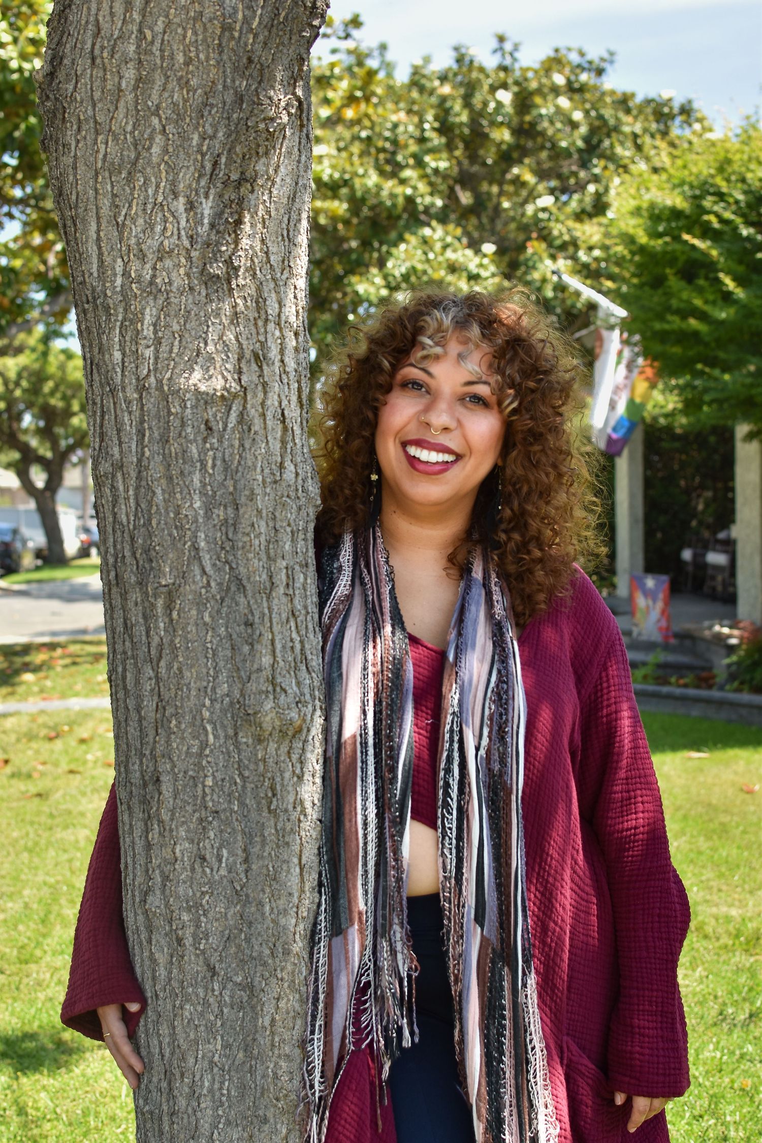 Person wearing a burgundy cardigan and striped scarf poses against a tree trunk in a sunny park setting.