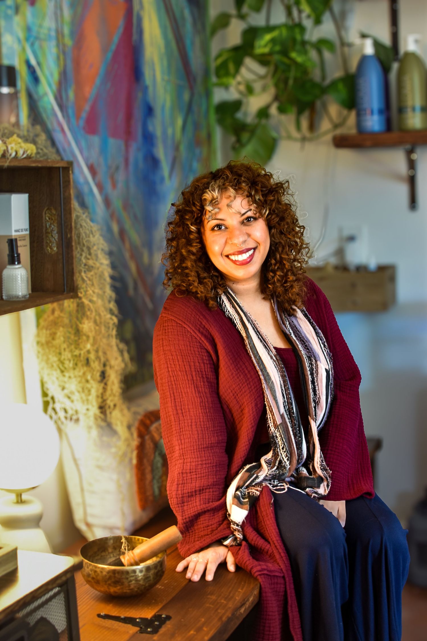 A person in a burgundy cardigan and printed scarf smiles warmly while standing in a rustic interior with shelves and decor.
