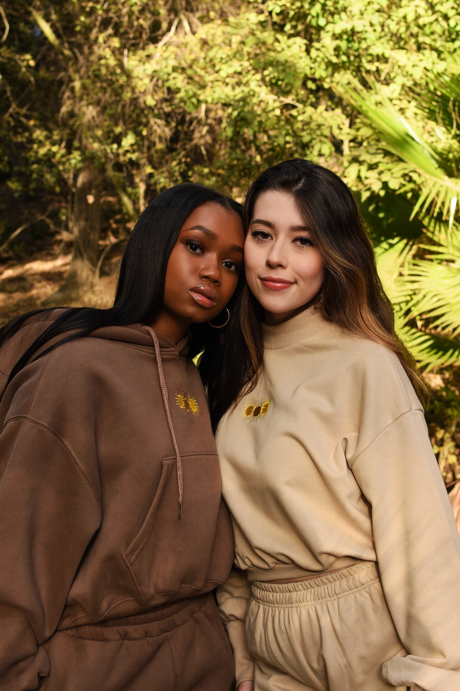 Two friends in matching neutral hoodies pose together in a sunny garden setting.