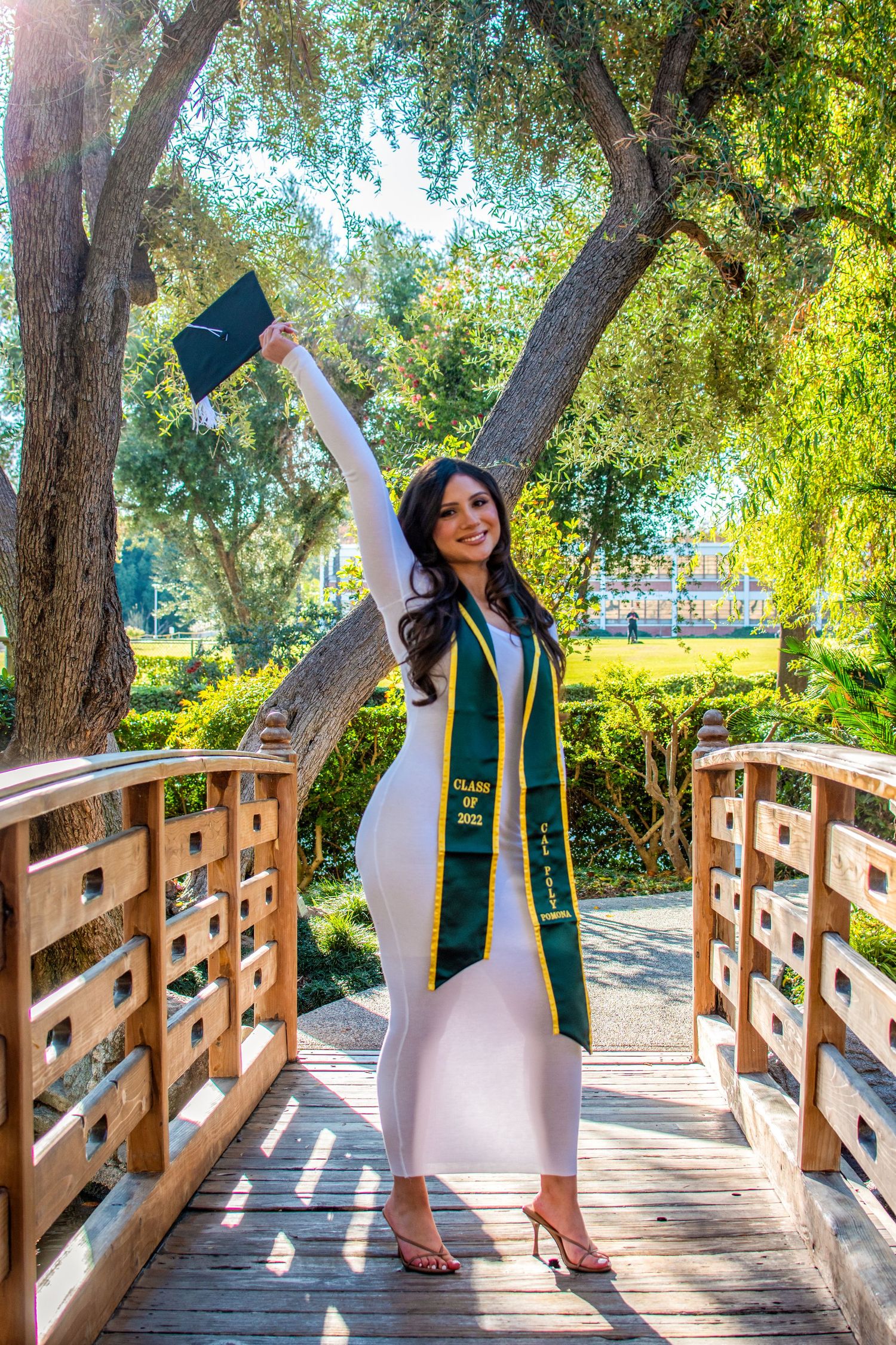 Excited graduate holding cap high celebrates on wooden bridge pathway surrounded by trees.