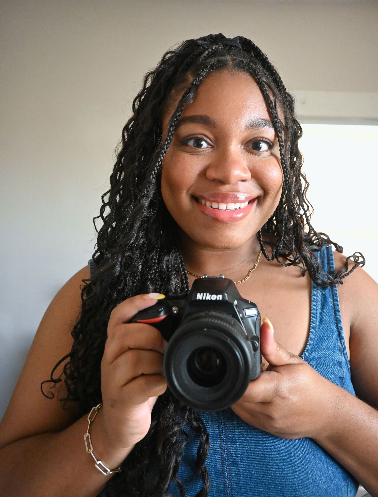 A creative photographer with braided hair holds a Nikon camera while wearing a blue tank top.