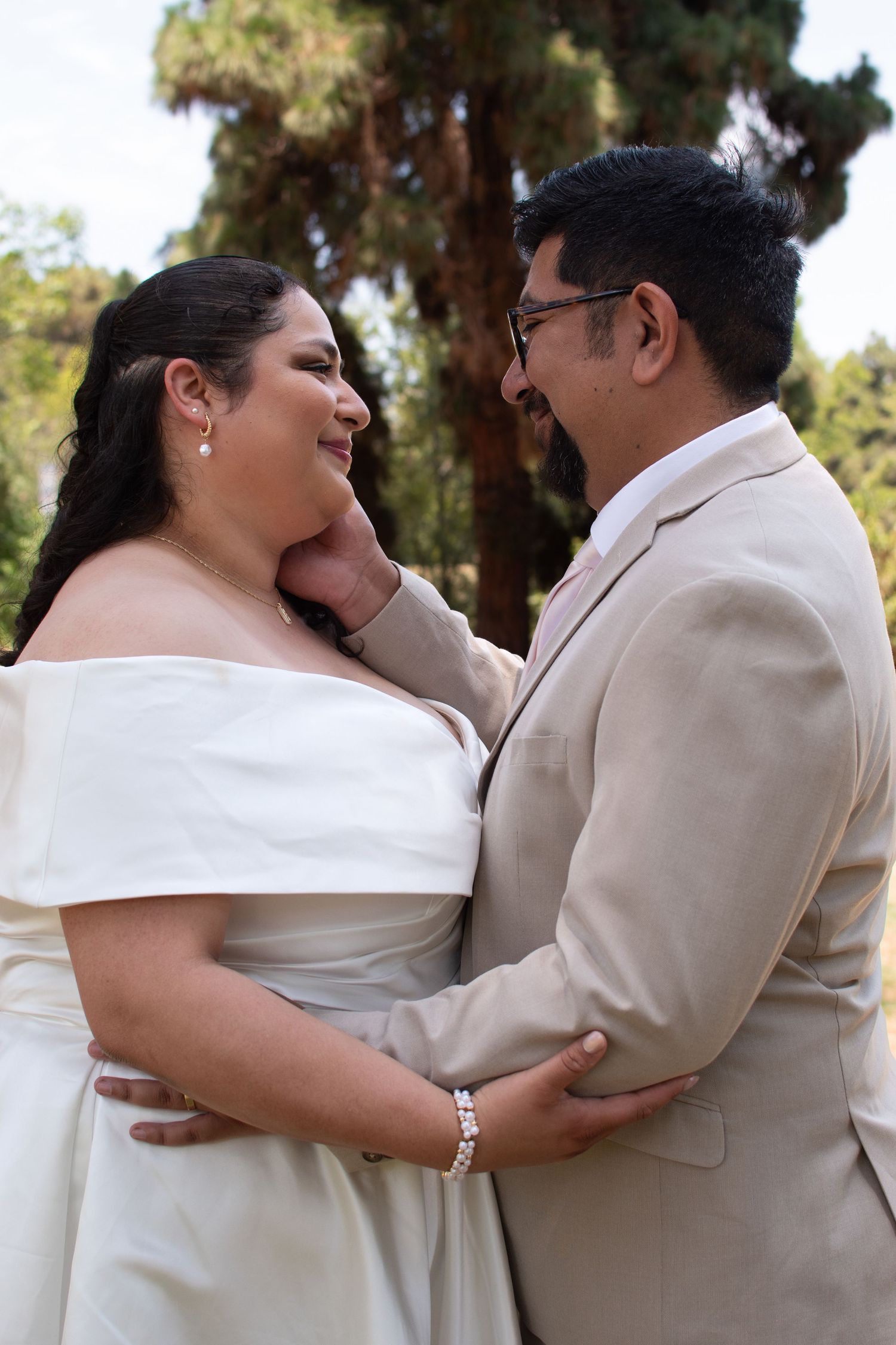 A happy couple in wedding attire sharing an intimate moment outdoors, with the bride in white and groom in beige.