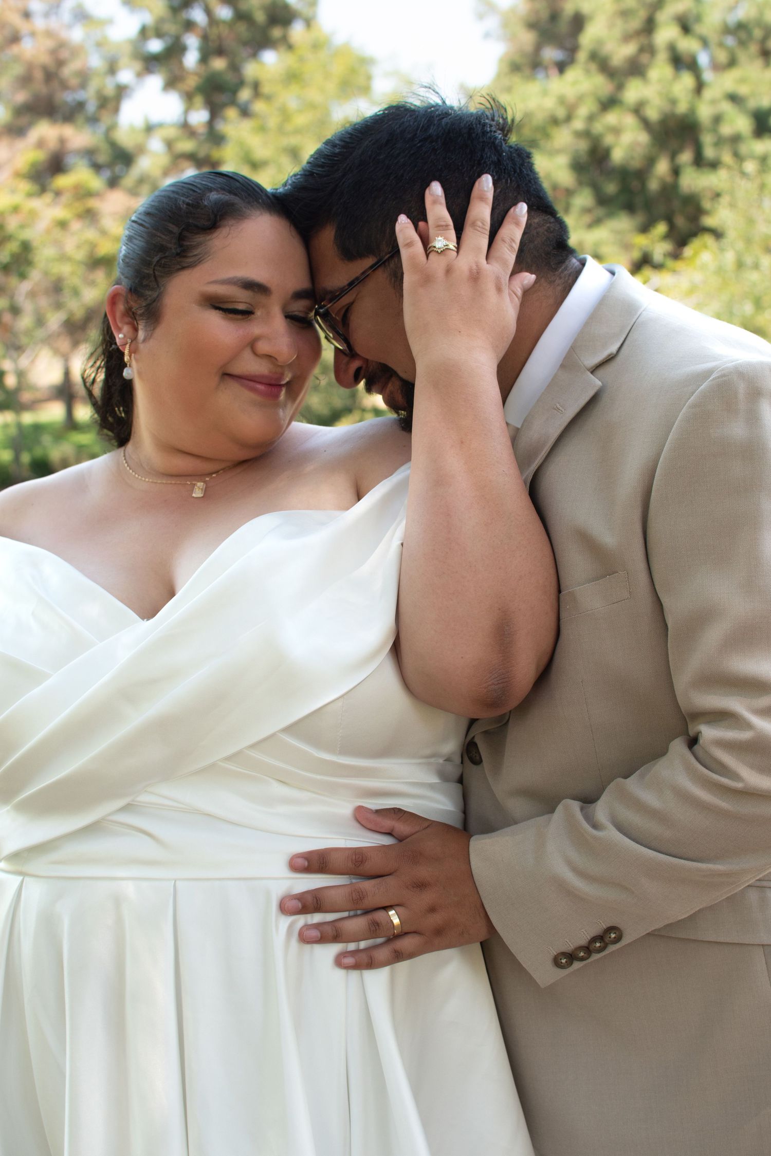 A romantic wedding photo showing a tender intimate moment between newlyweds in white dress and beige suit outdoors.