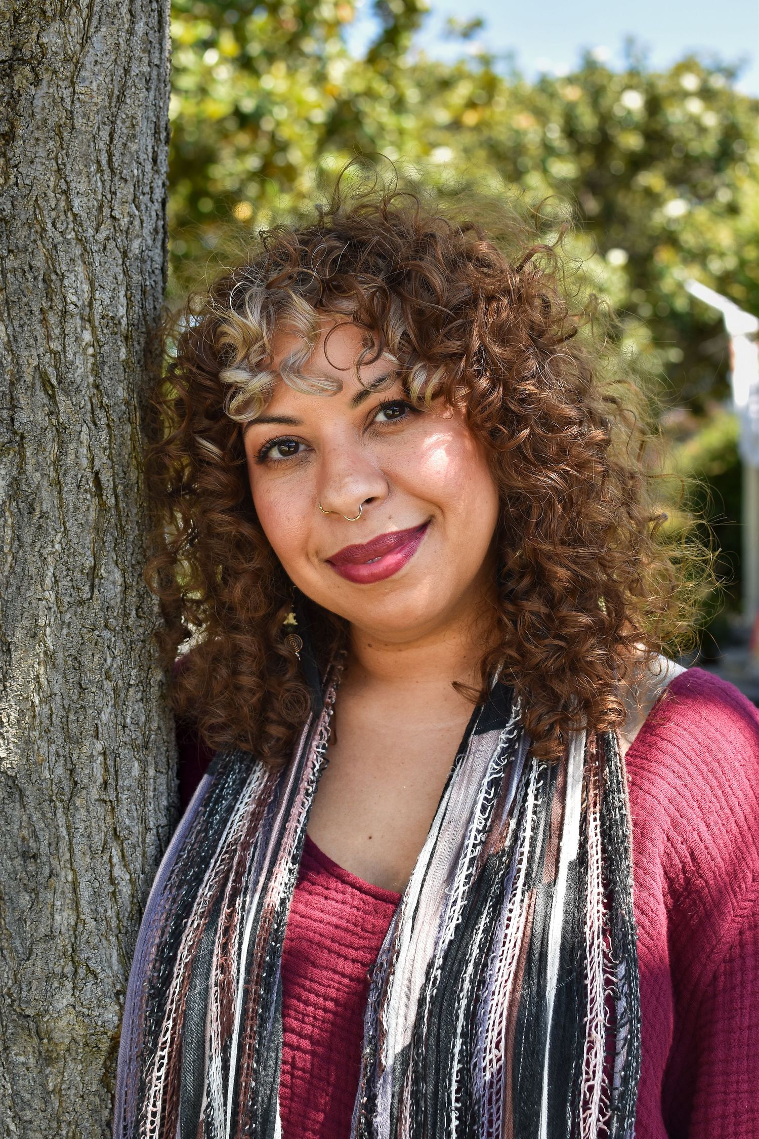 A person with curly hair smiles while leaning against a tree trunk wearing a red sweater and striped scarf outdoors.