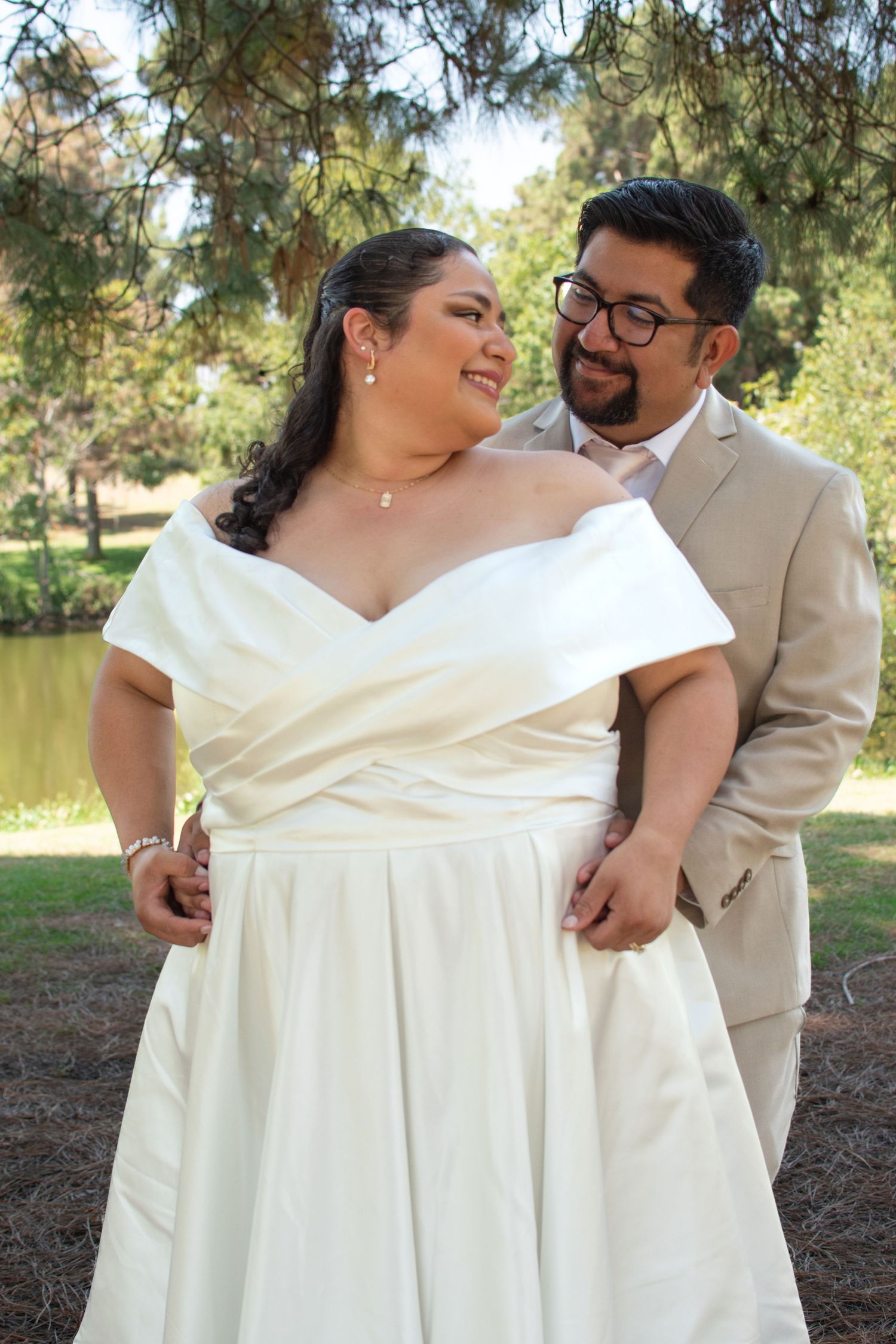 A romantic outdoor wedding portrait of a couple in white dress and beige suit standing by a lake on a sunny day.