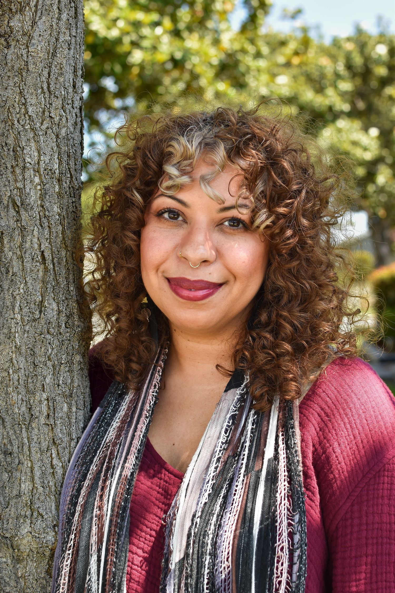 Close-up portrait outdoors showing curly hair and striped scarf against green trees in background.
