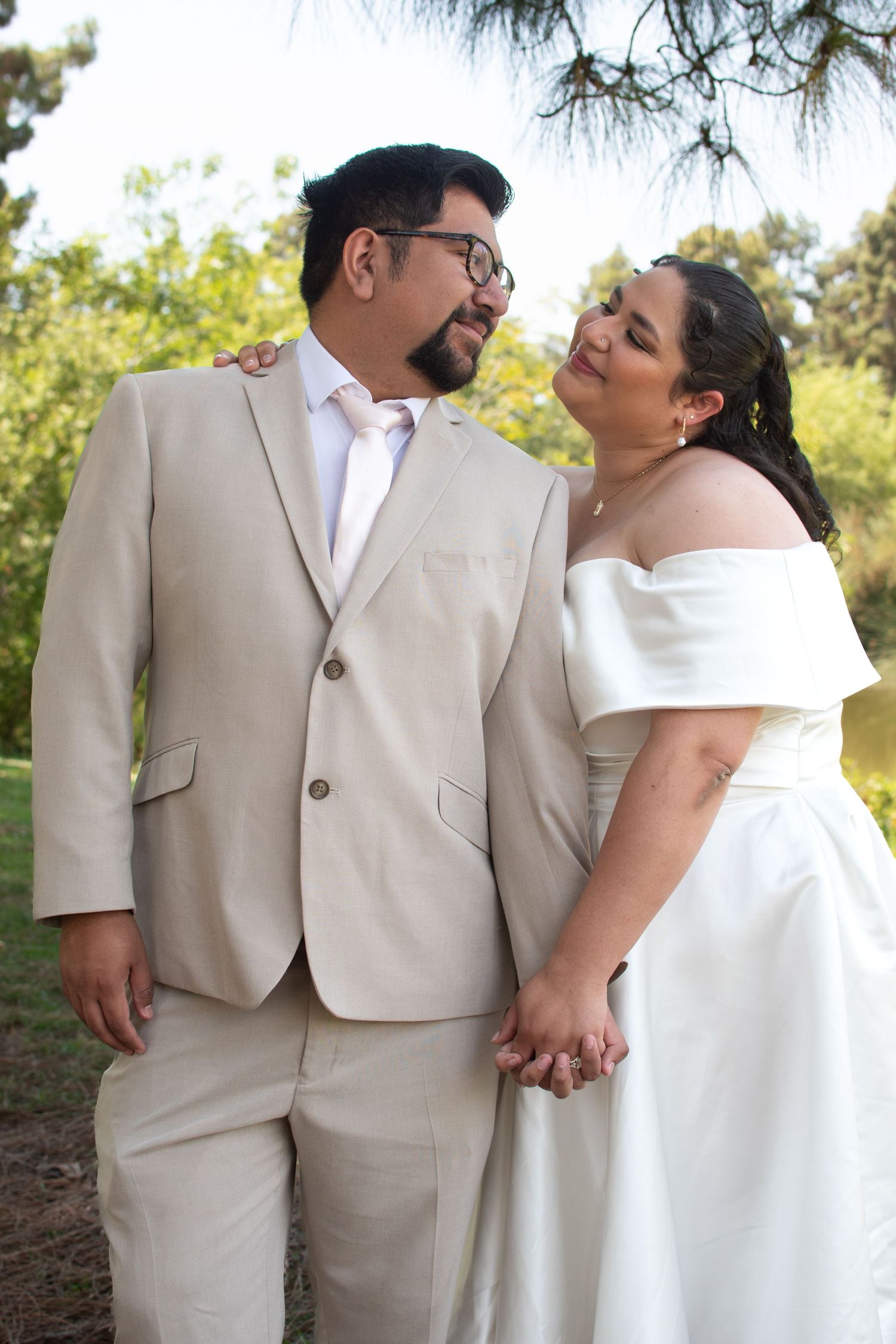 A couple shares an intimate moment in wedding attire with the bride in white and groom in beige against a garden backdrop.