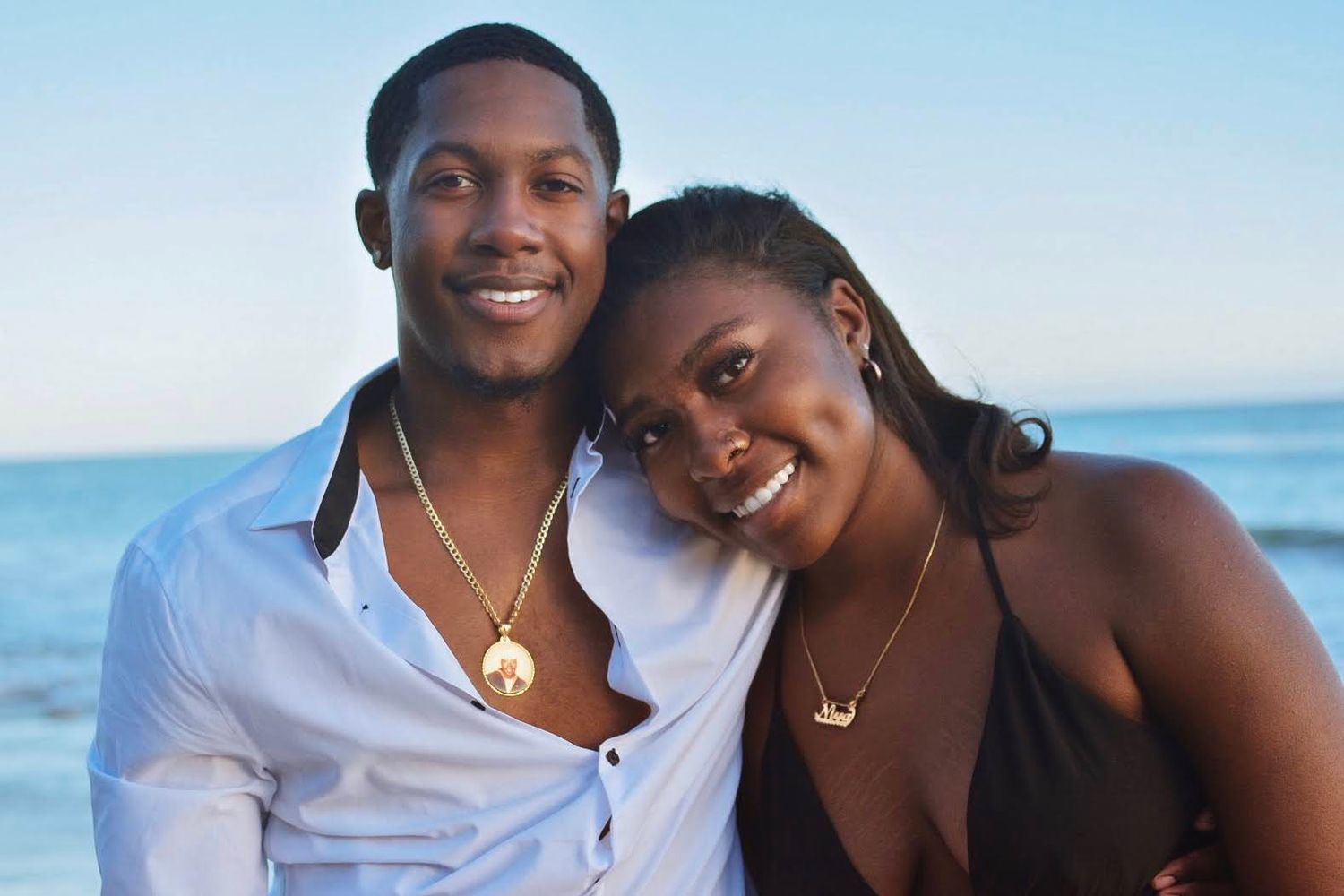 Happy couple embraces while posing together on sunny beach with ocean in background.