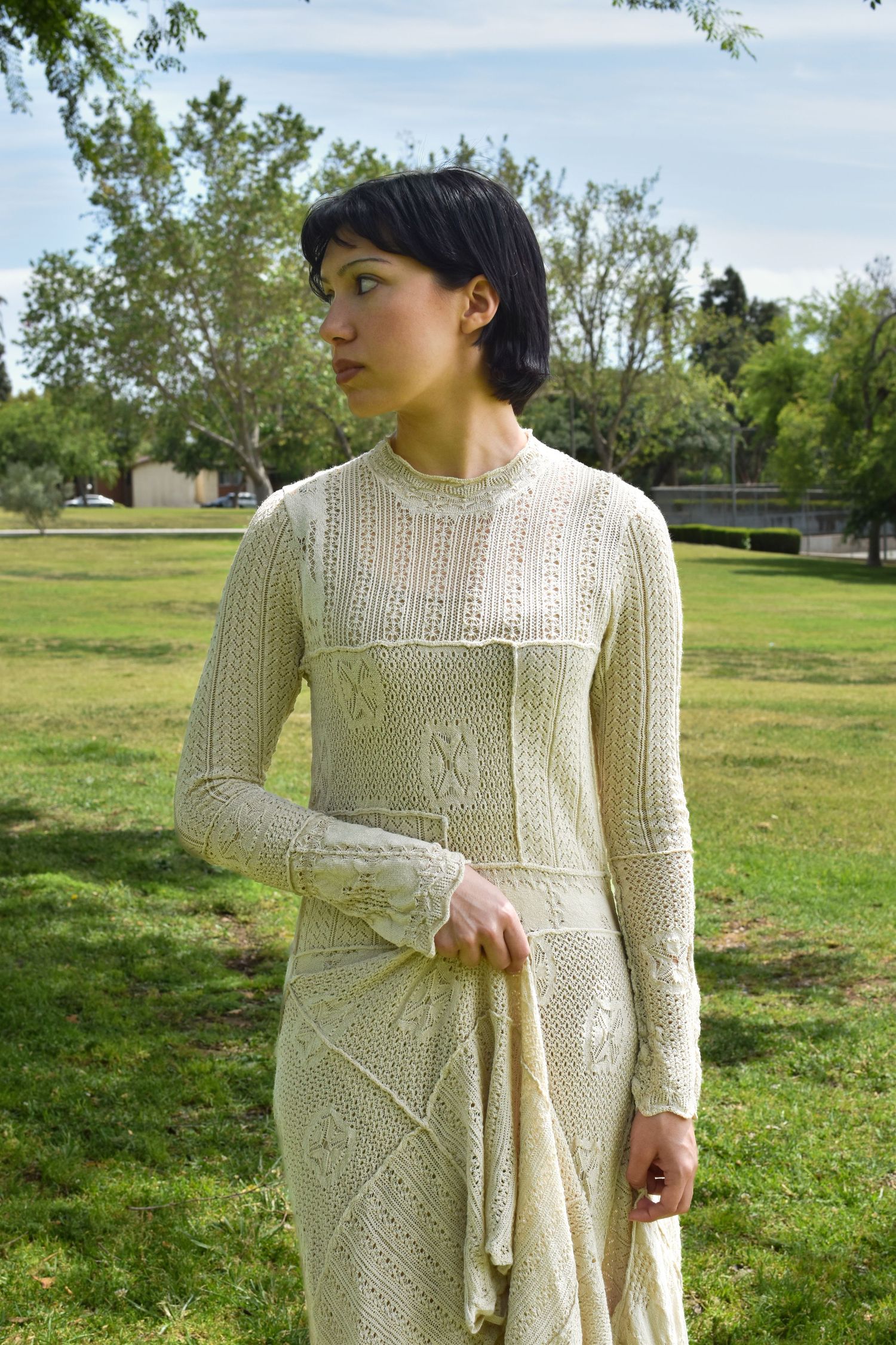 A person wearing a detailed cream knitted dress stands in a sunlit park setting.