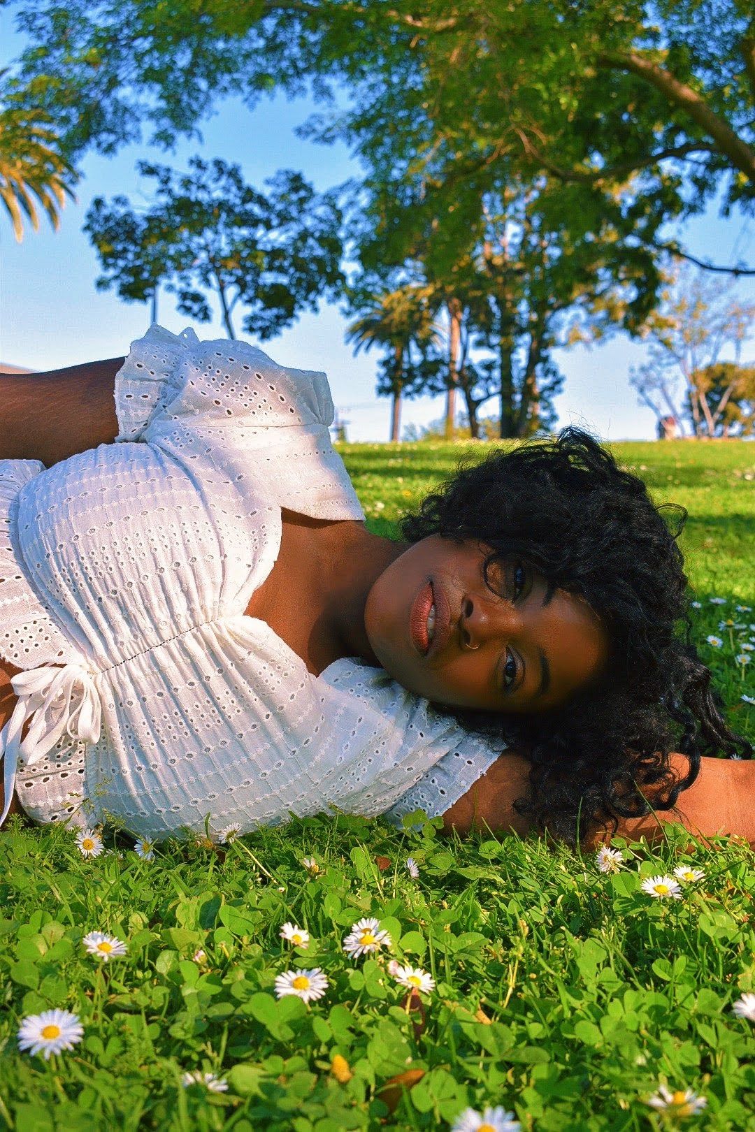 A person in a white dress lying on grass with small daisies on a sunny day.
