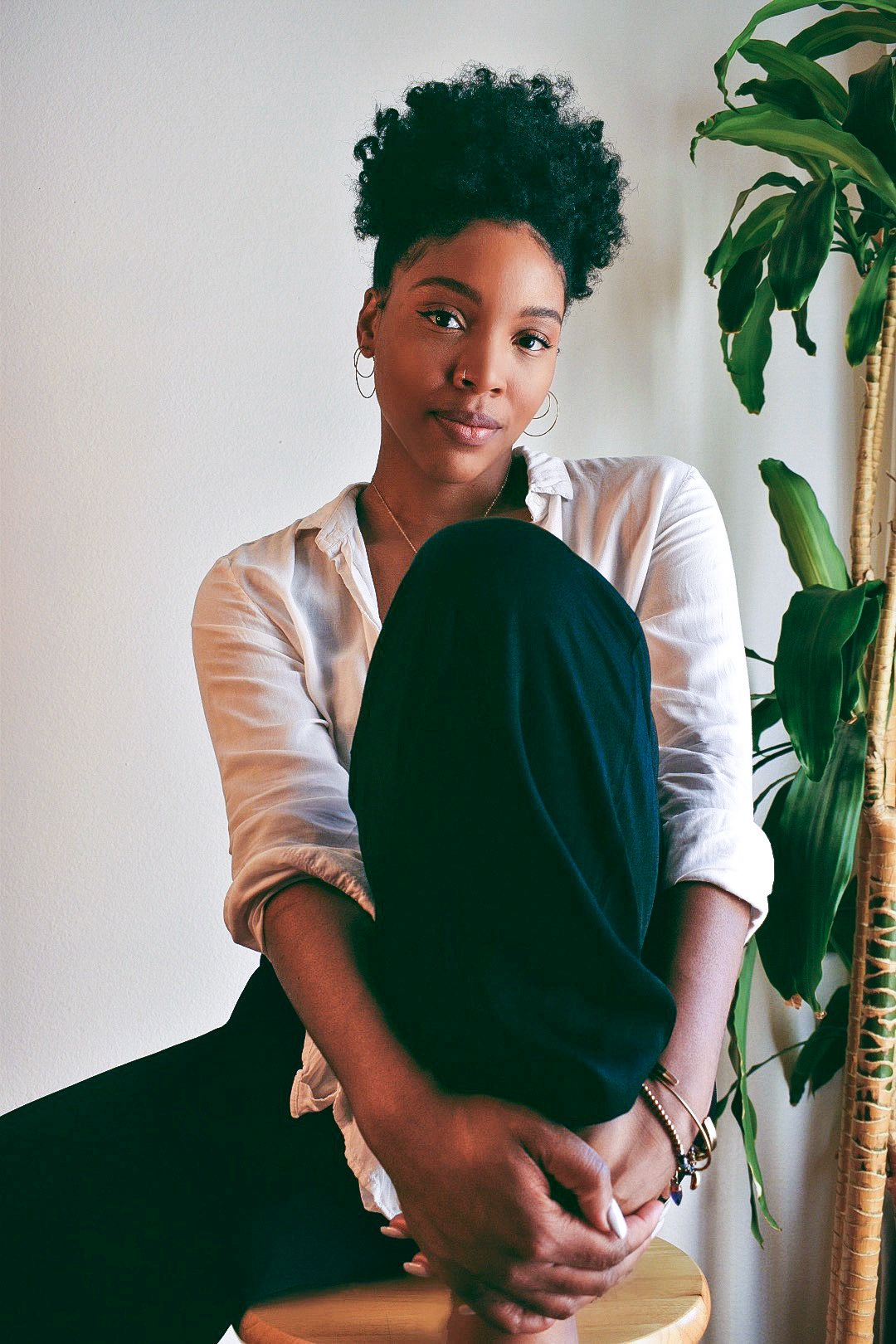 A person in a white button-up shirt and dark pants sits casually beside a large potted plant against a white wall.