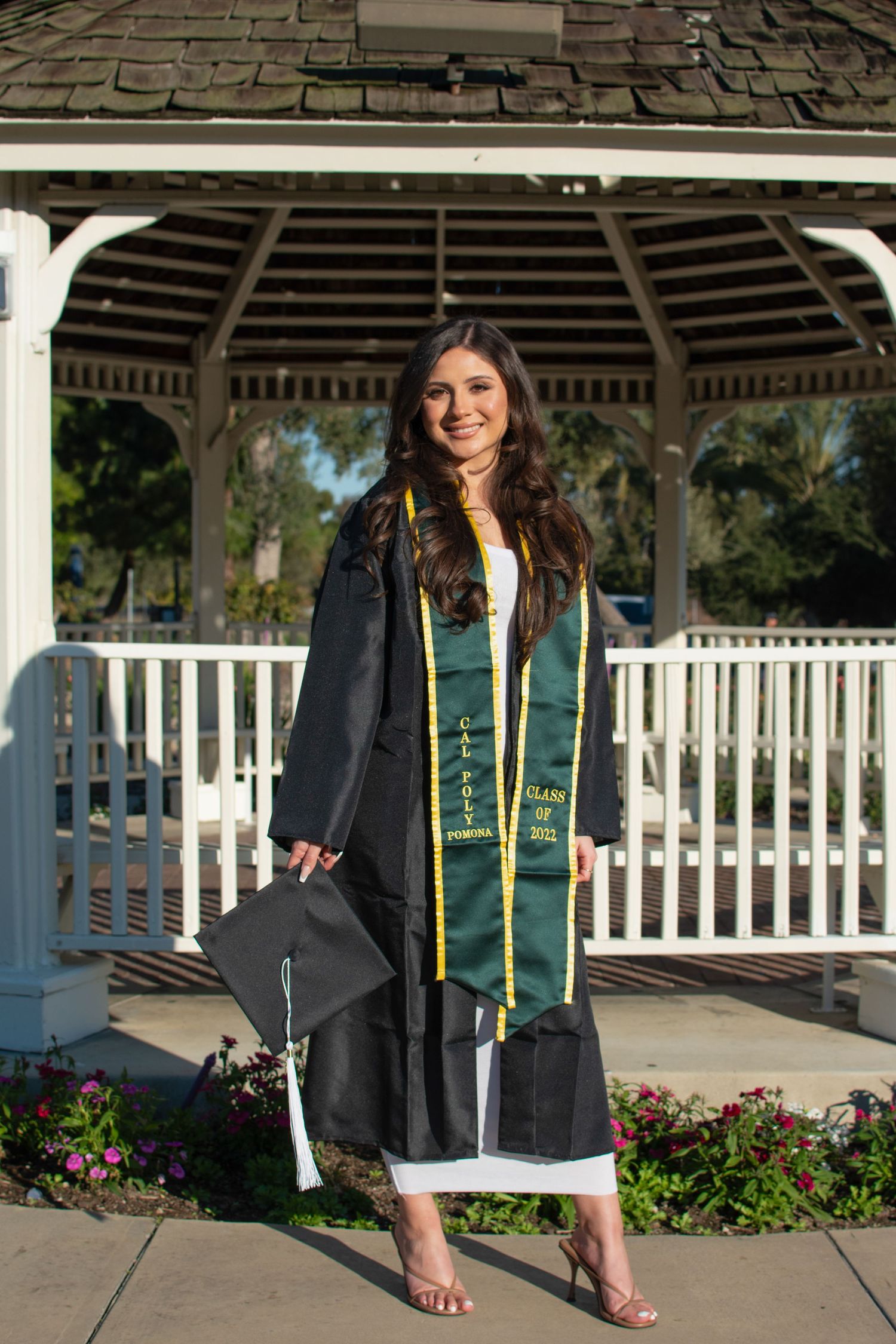 Graduate in traditional black cap and gown poses in front of white gazebo with diploma and graduation stole.