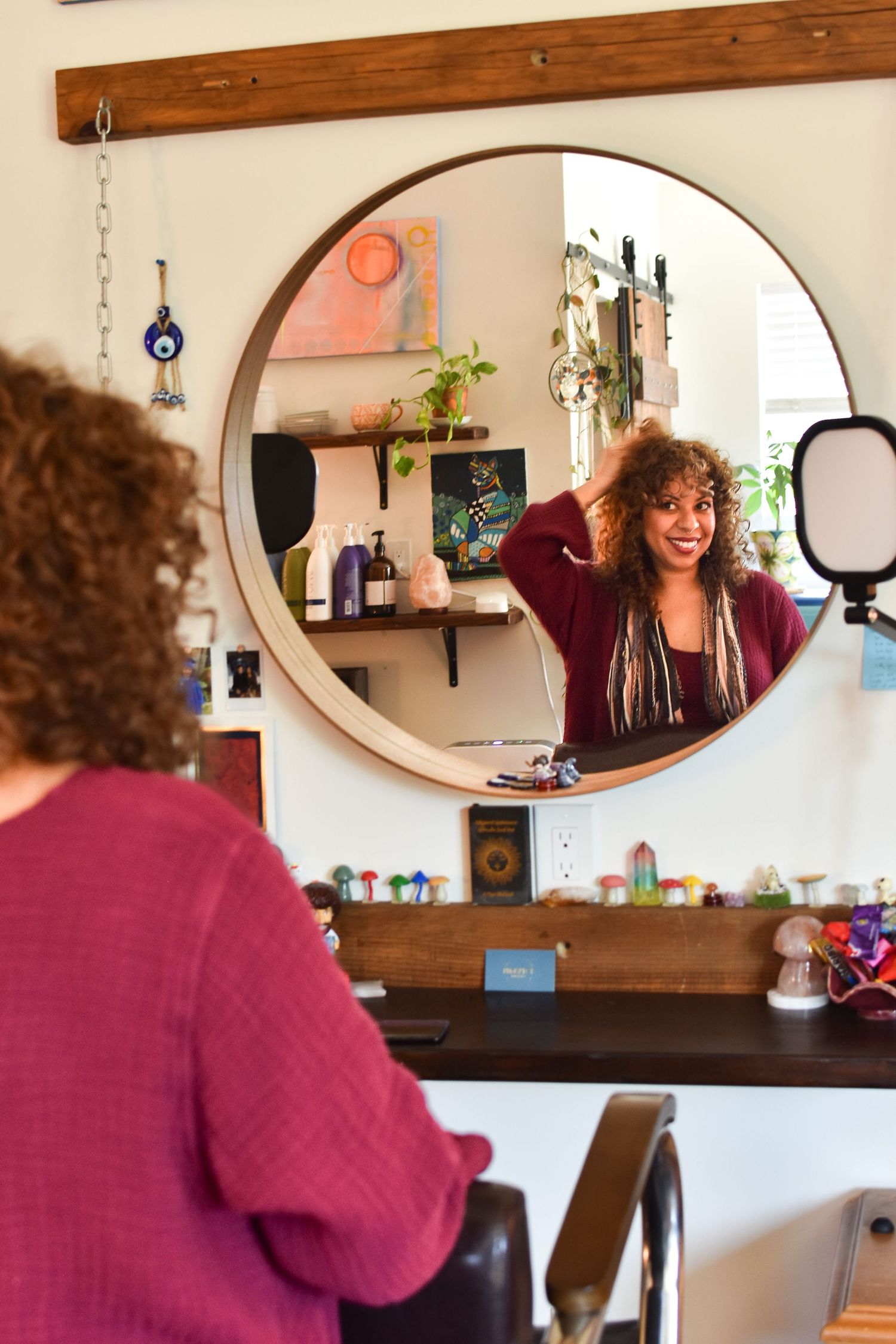 A round mirror on a wooden wall reflects colorful decorative items and a ring light in a salon setting.