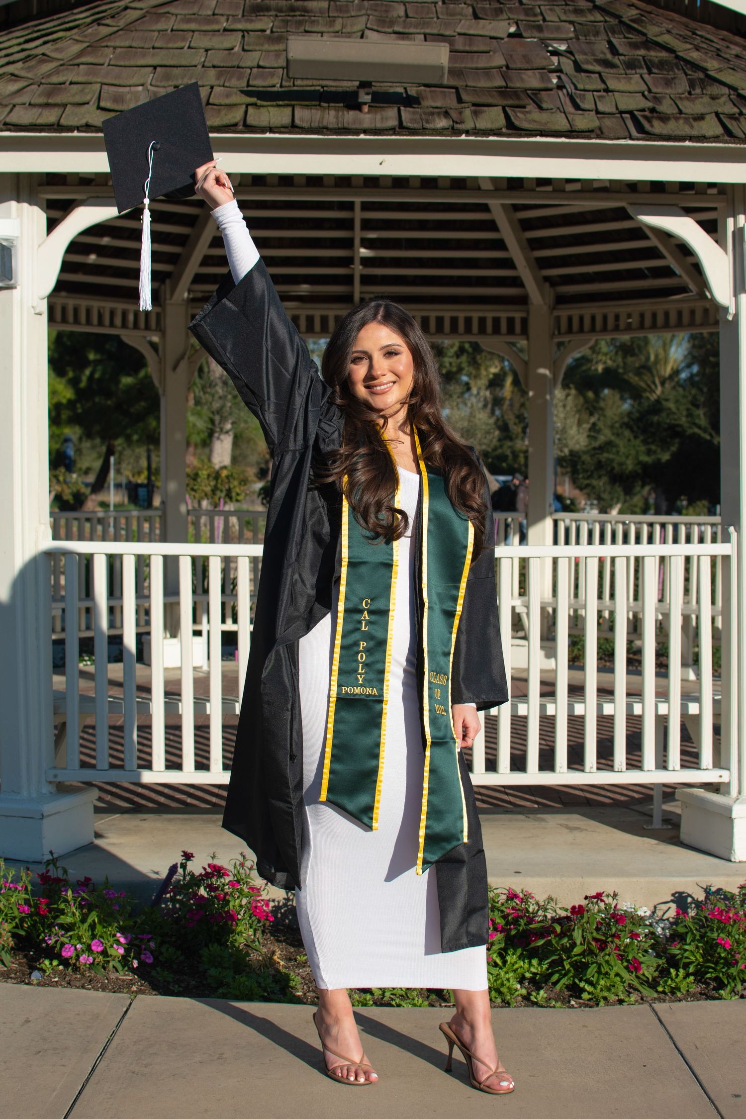 Graduate in black graduation gown raises cap victoriously while posing in front of white gazebo.