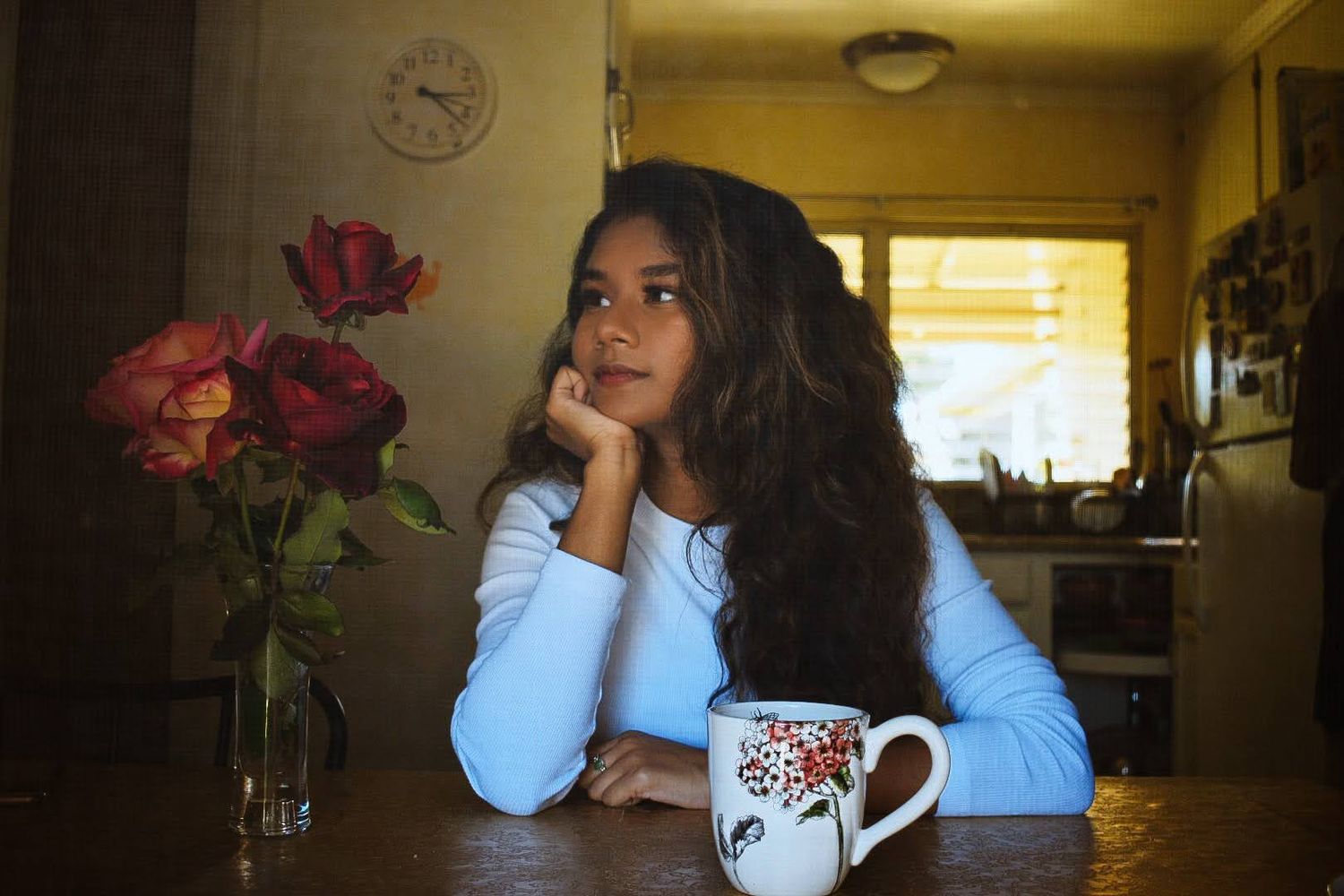 Person posing thoughtfully at table with decorative mug and roses in warm indoor lighting.